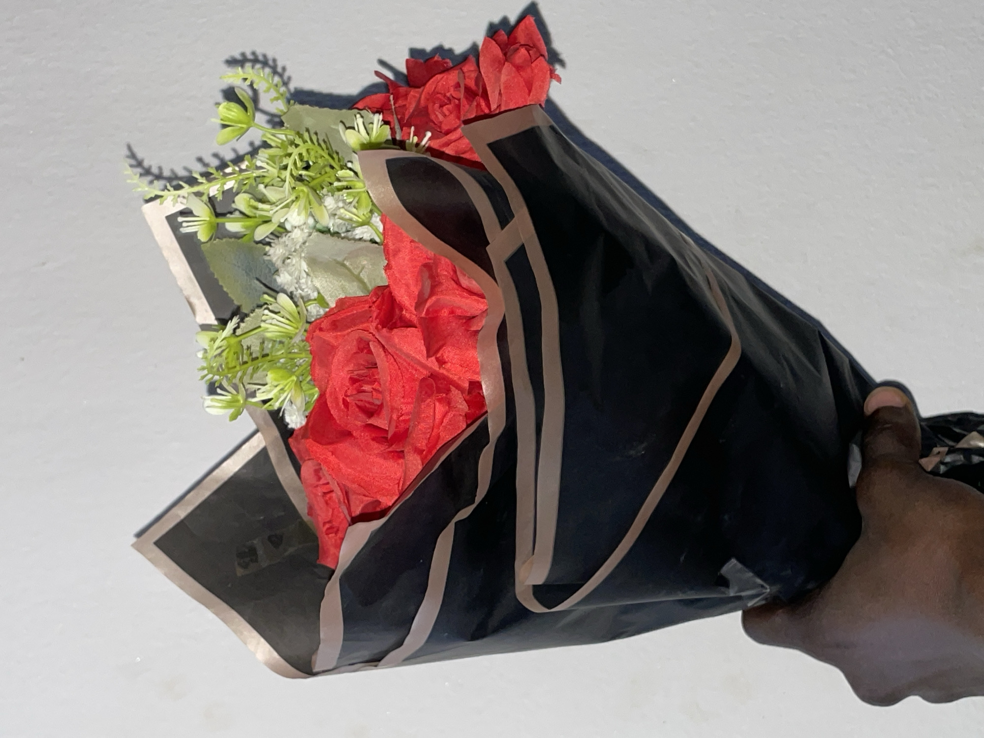 A hand holds a bouquet of red roses, partially wrapped in black paper with brown trim. Small white and green accent flowers are interspersed with the roses. The background is a textured, light-colored wall. The image appears to be taken indoors. The specific location within Durimi, Nigeria cannot be determined from the visual information provided.