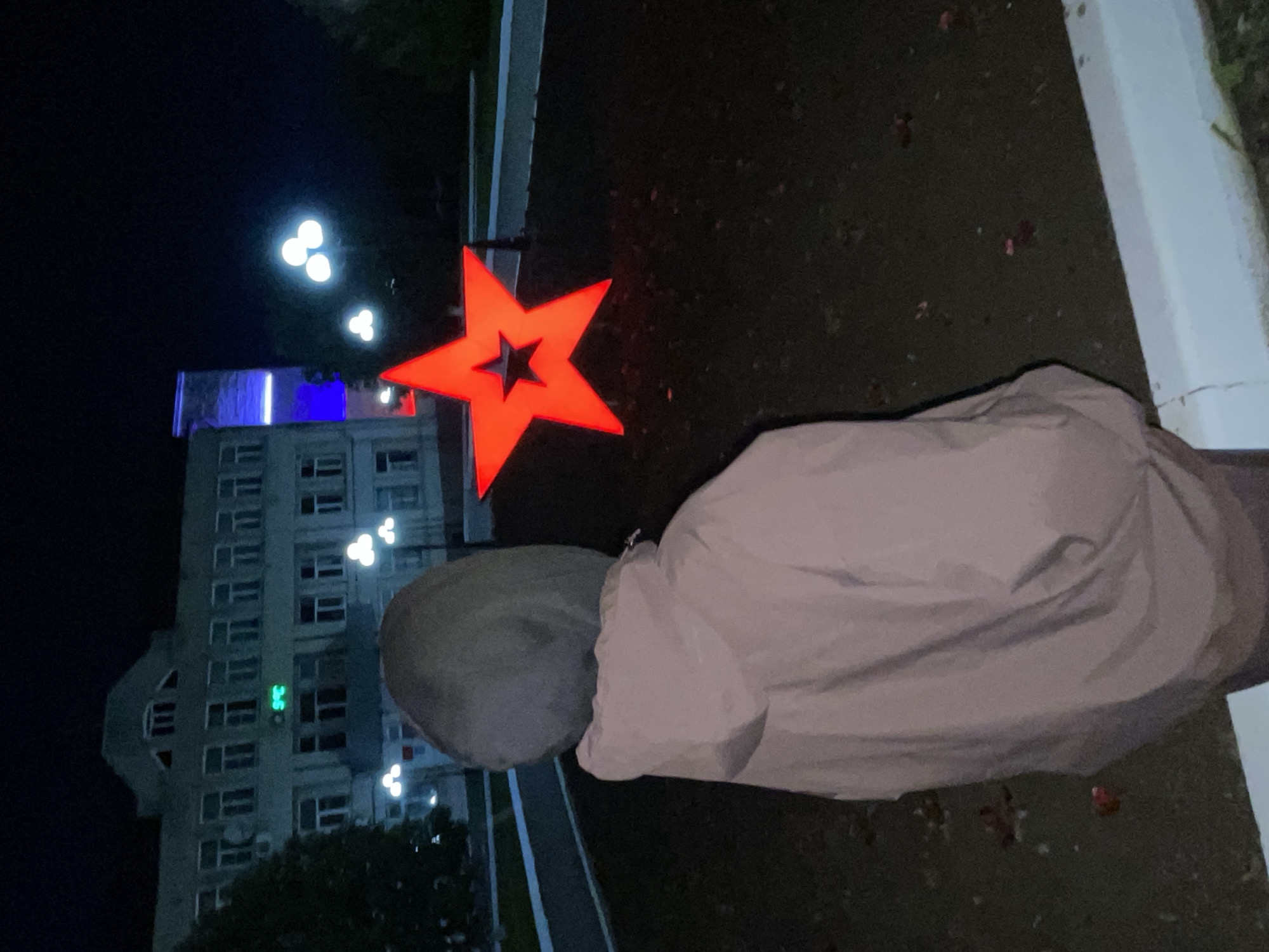 This media file depicts an outdoor night scene in Красные Баки, Russia, observed from behind a person.
**Subjects:**
The primary subject is a person, seen from the back, wearing a light-colored, possibly beige or grey, hooded jacket. Their head is covered by the hood. In front of the person, a large, glowing red five-pointed star is prominently displayed, featuring a smaller, dark star shape cut out from its center. Further into the background stands a tall, multi-story building with numerous windows, constructed from light-colored concrete. Several streetlights, emitting bright white light, are scattered throughout the scene.
**Activity or event taking place:**
The person appears to be standing or slowly walking, observing the illuminated star and the building. No specific action or event is discernible beyond this observation.
**Notable details:**
The time of day is clearly night, indicated by the dark sky and the reliance on artificial light sources. The red star glows intensely, dominating the mid-ground. The streetlights provide circular pools of white light. A distinct blue light illuminates a section at the top of the building. On one of the building's upper floors, a green digital display is visible, showing what appears to be the numbers "3.5" or similar characters. The ground is dark, likely paved, with some small, indistinct reddish specks. The building's architecture suggests a functional, possibly older, style. The overall mood is quiet and contemplative, characteristic of an urban evening.