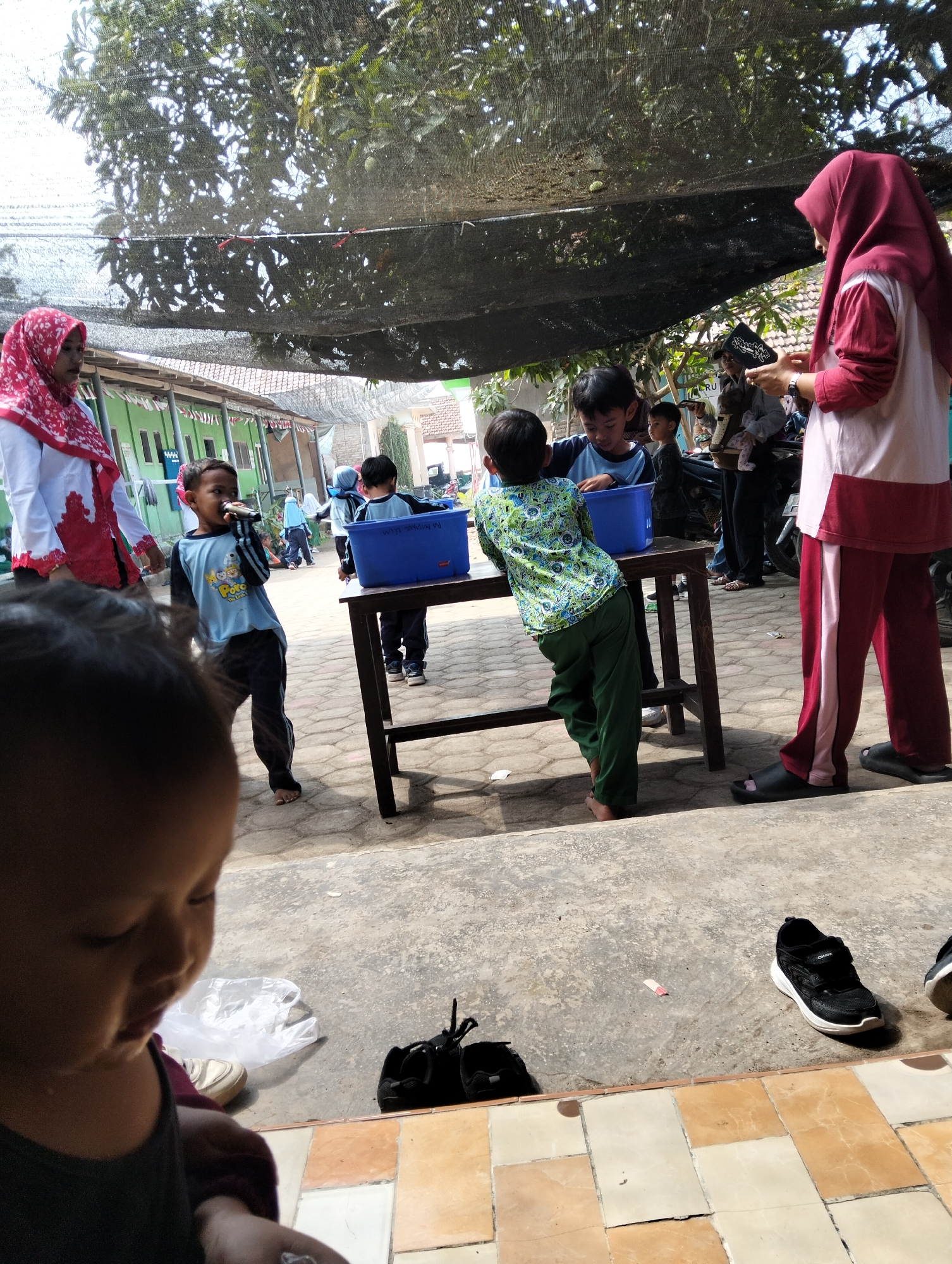 In Desa Wringin Putih, Indonesia, a group of children are gathered outdoors, possibly at a school or community event, near a building with green walls and red and white flags hanging. A dark mesh shade is stretched overhead, with trees visible through it. In the foreground, a young child's face is partially visible. Several other children are interacting around a wooden table with blue plastic tubs on it. One boy in a blue and yellow shirt is holding a microphone. Another boy in a green patterned shirt and green pants is leaning over the table. Two other boys, one in a blue shirt and another in a darker shirt, are also near the table. A woman in a red hijab and a white and red top is standing to the right, while another woman in a red hijab and a white and red top is on the left. Several pairs of shoes are on the ground.