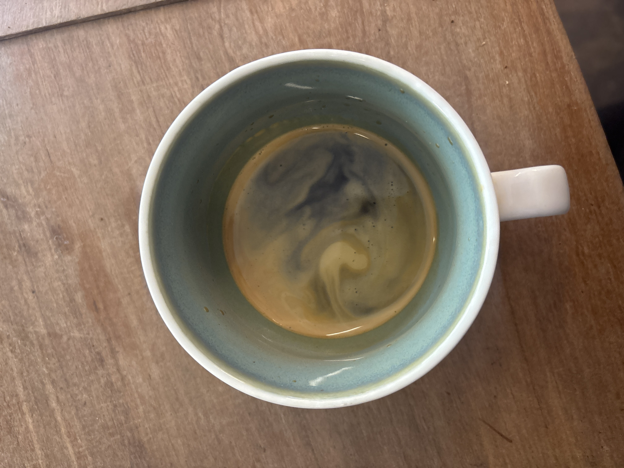 An overhead shot presents a single ceramic mug resting on a light-brown wooden surface. The mug's interior is a gradient of light blue to muted teal at the rim, contrasting with its white exterior and a small, white handle visible on the right side. Inside the mug, a dark liquid, likely espresso or strong coffee, is visible, topped with a rich, golden-brown crema. Distinct darker swirls are present within the crema and liquid, suggesting recent pouring or gentle agitation. The wooden surface displays a clear grain and some subtle imperfections. The lighting is soft and even, casting no harsh shadows. The image depicts a tranquil scene, commonly associated with a coffee break. No individuals are present, and there is no visible text. While the location context is provided as Buenos Aires, Argentina, the image itself does not contain any specific visual cues to identify this location.