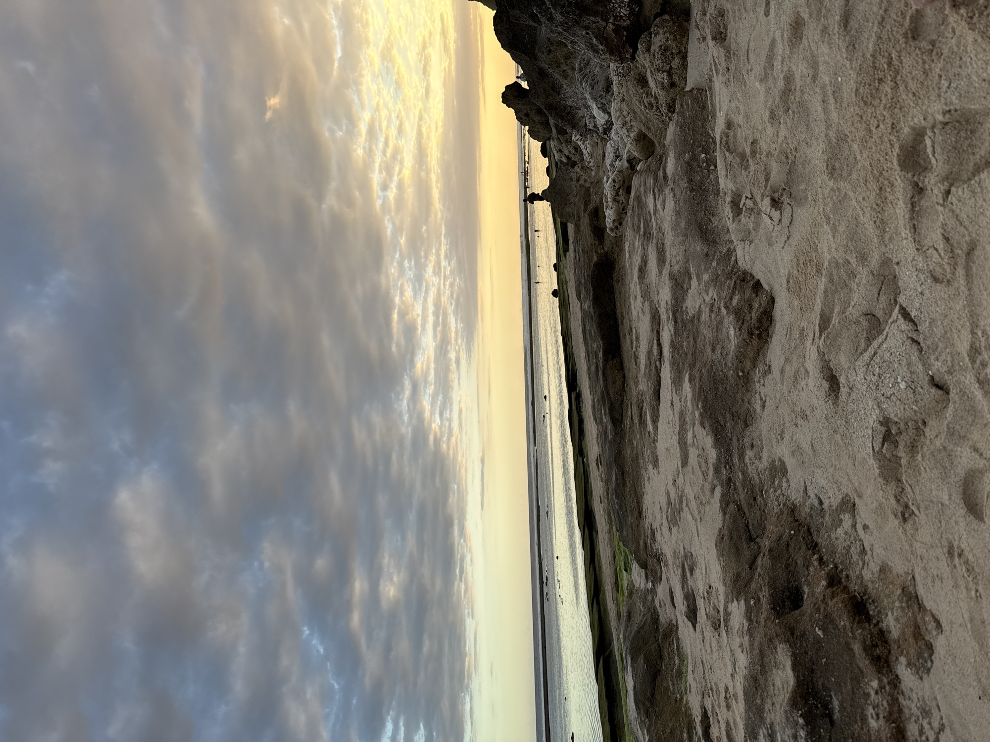 The image depicts a beach scene in Kuta Selatan, Indonesia. On the right is a rocky, sandy cliff face with visible erosion patterns. The sand is a light tan color with darker patches, possibly due to moisture or different sediment composition. To the left of the cliff face, there's a flat beach area, possibly tidal flats, with standing water reflecting the sky. The horizon line is at the center of the image. People are visible as tiny figures in the distance on the beach. The sky is filled with clouds, ranging from shades of gray to light yellow, suggesting either sunrise or sunset. The lighting is soft and diffused, creating a calm, tranquil ambiance.
