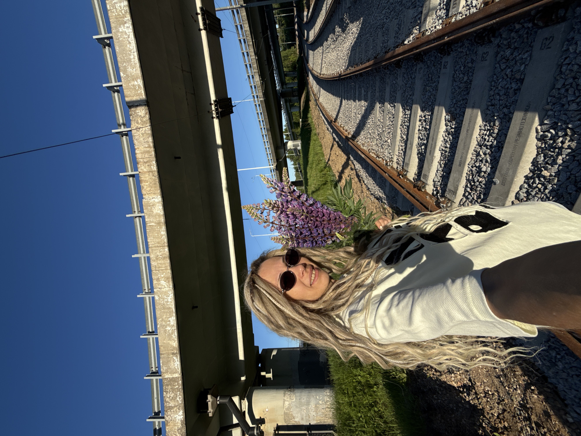 An adult female, appearing to be in her 30s or 40s, with long, blonde dreadlock-style hair and sunglasses, smiles at the camera while taking a selfie. She wears a white long-sleeved top with black abstract or geometric designs. In her left arm, she holds a bouquet of purple lupine flowers. She stands outdoors on the left side of the frame, positioned beside railway tracks.
The scene features multiple concrete overpass bridges against a clear blue sky. A large concrete bridge structure extends overhead, under which the woman stands. This bridge is supported by concrete pillars on the left, and electrical or signal lines are suspended from its underside. Another, narrower concrete bridge with metal guardrails is visible further back and higher up, spanning the scene.
Several sets of railway tracks, featuring rusty metal rails on grey gravel ballast and concrete sleepers, curve to the right into the distance. In the foreground, text visible on some concrete sleepers reads "ООО ПКО «ЕВРОХИМ РЕЗИНОТЕХНИКА»" along with alphanumeric codes like "R3" and "02". Green grass and other vegetation grow on the embankments and around the bridge pillars. Strong sunlight from the right casts distinct shadows across the railway tracks. This scene is located in Rusanovo, Russia.