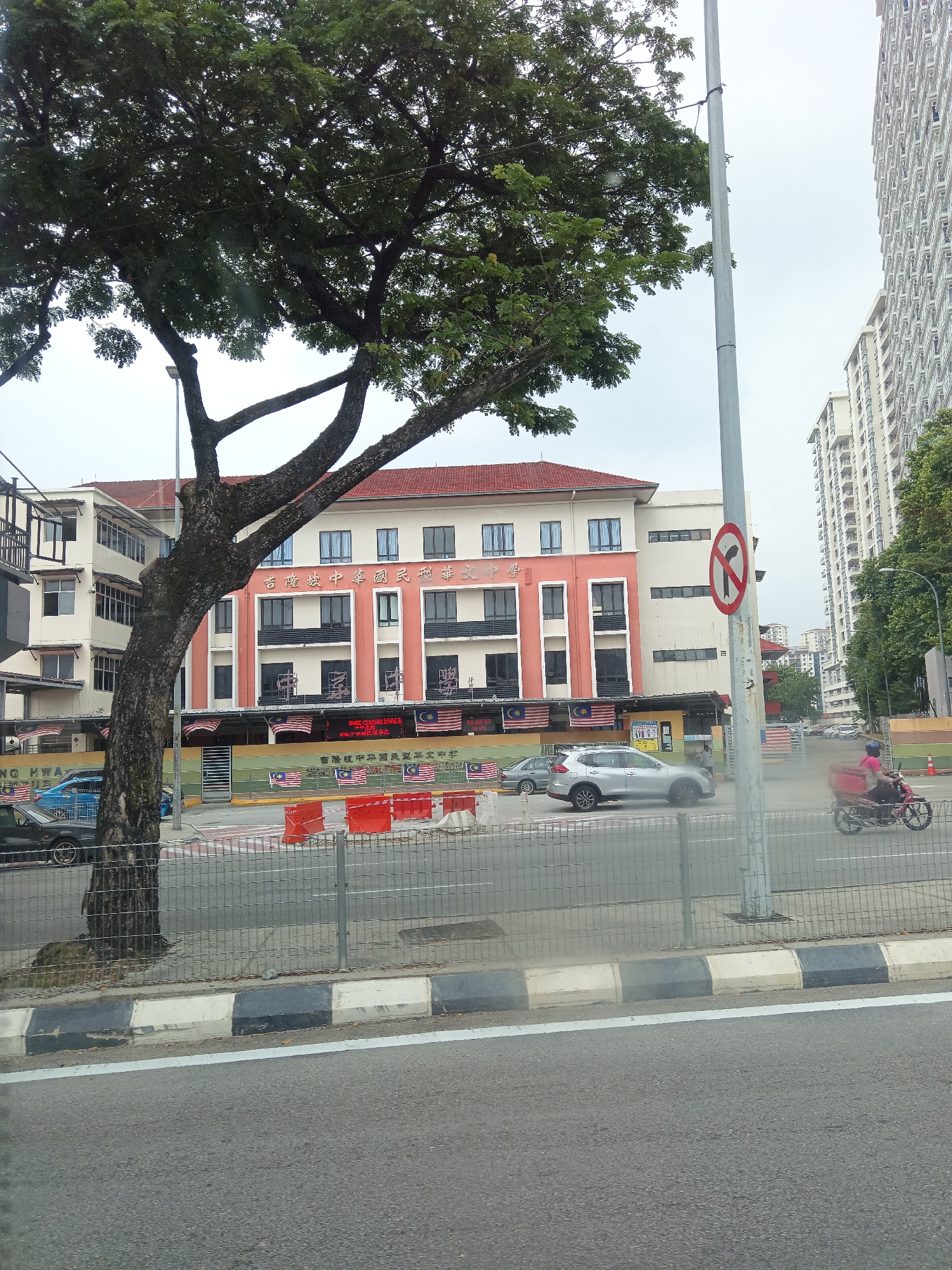 The image captures a street scene in Kuala Lumpur, Malaysia. In the foreground, a road with a white line and a curb marked with alternating black and white segments is visible. A metal fence runs parallel to the road. Beyond the fence, a tree with a thick trunk and spreading branches occupies the left side of the frame, partially obscuring a building. The building is a multi-story structure with a red tiled roof and a facade featuring cream and orange sections with numerous windows. Malaysian flags are displayed along the front of the building.
Several cars are parked or driving on the road. To the right of the building, a tall, modern white building with many windows dominates the background. A lamppost stands near the right edge of the frame, displaying a circular red and white sign with a black arrow indicating a no left turn. Further down the road, a person is riding a red motorcycle with a red box attached to the back. The sky is overcast.
