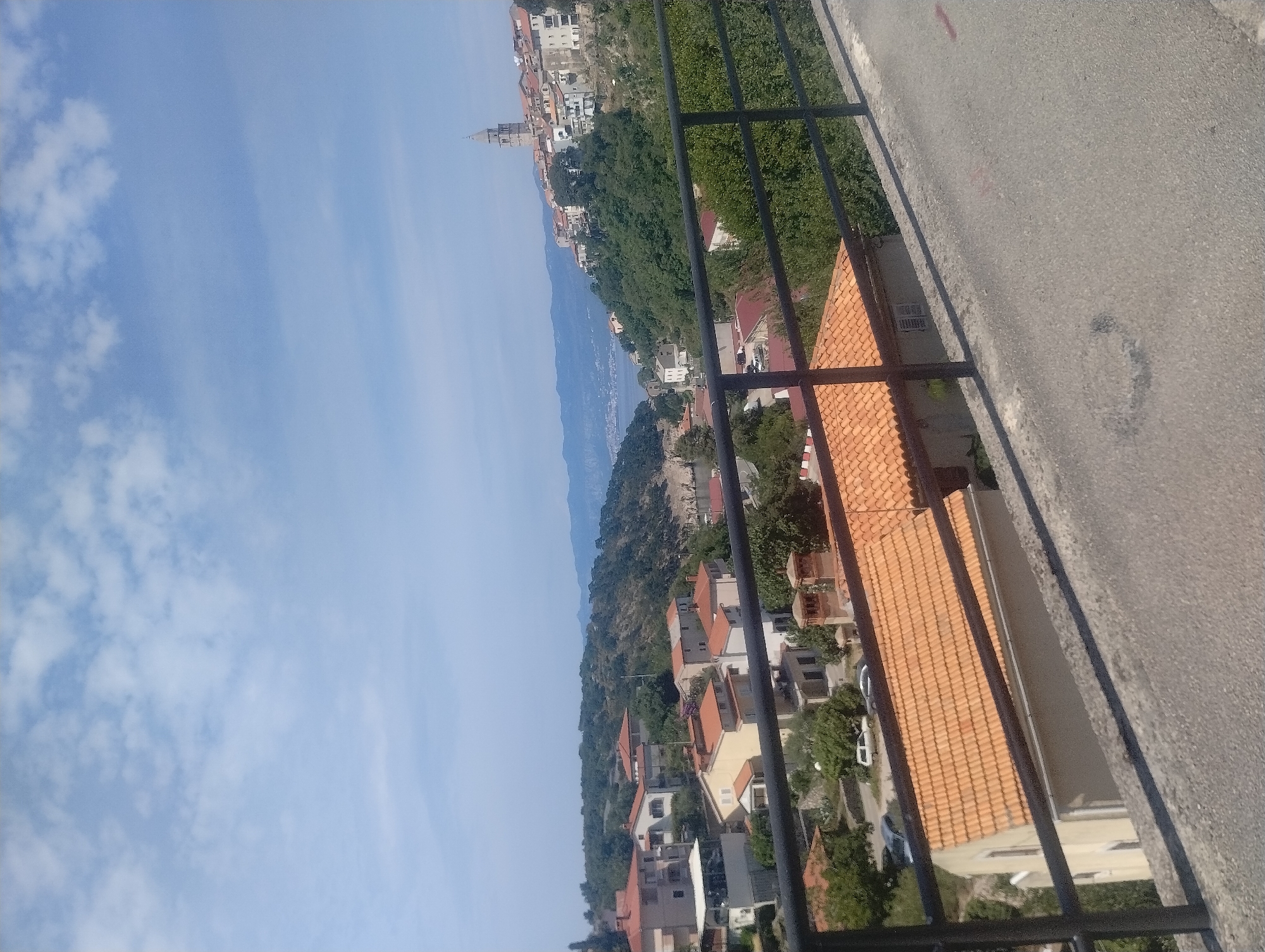 A daytime, elevated, wide shot captures a coastal town on a hillside under a partly cloudy blue sky. In the foreground, a gray asphalt road curves from the right, bordered by a black metal railing that extends across the frame. Beyond the railing, several houses with orange-tiled roofs are visible. Some houses are closer to the foreground, with their details clearly defined, while others are further back, becoming less distinct.
The middle ground shows a densely wooded hillside with more houses interspersed among the trees. The town extends up the hill, with buildings clustered together, many featuring the characteristic orange tile roofs. A prominent bell tower rises above the town, indicating a church or cathedral.
In the background, a body of water, likely the Adriatic Sea, stretches to the horizon, with distant, hazy mountains framing the view. The sky is a bright blue, with scattered white clouds that cast soft shadows. The overall impression is of a sunny, picturesque Mediterranean town.