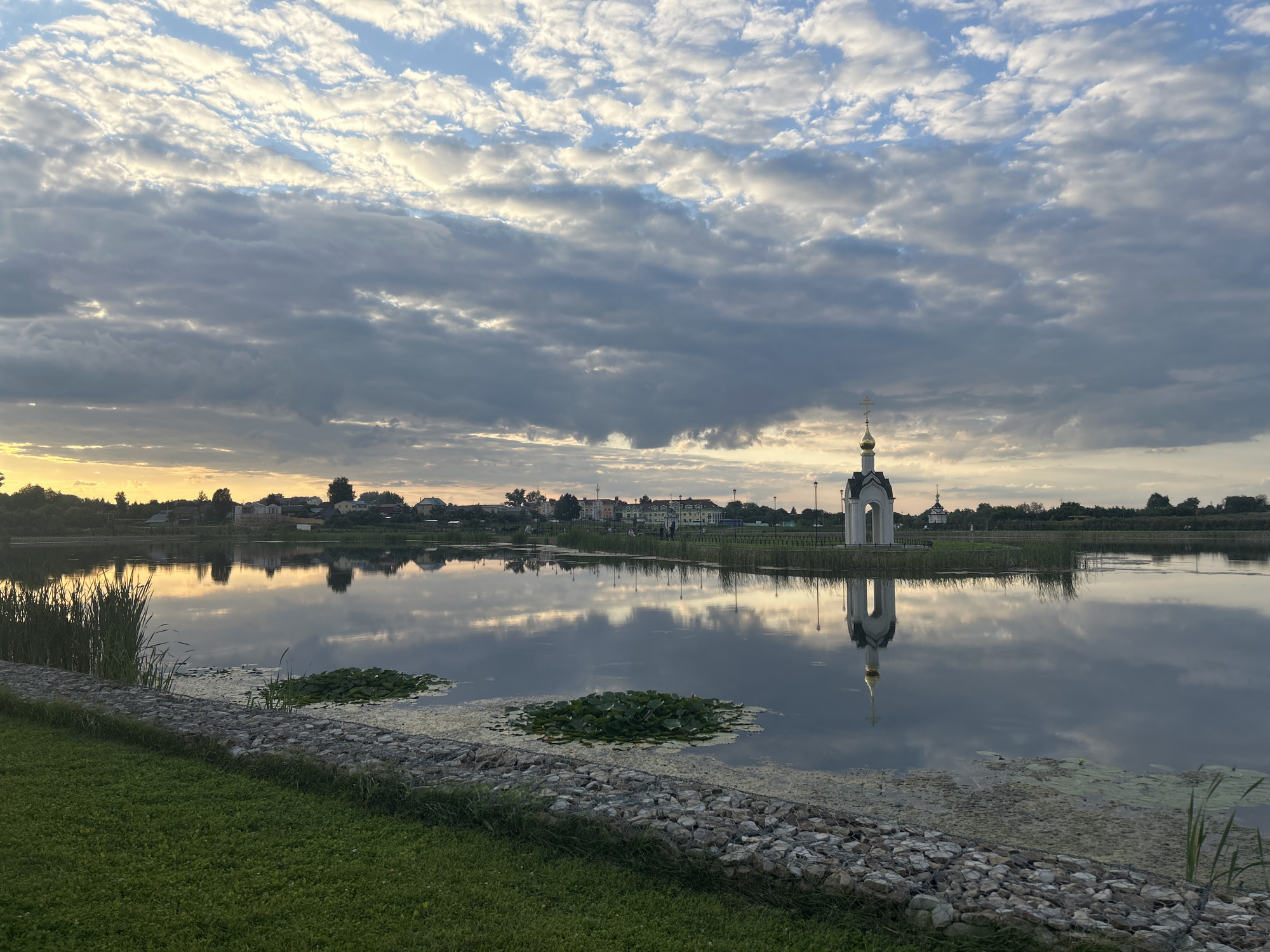 The image shows a serene lake in Diveyevo, Russia, during sunset. The sky is filled with dramatic clouds, illuminated by the warm hues of the setting sun, creating a breathtaking reflection on the calm water. On the far bank of the lake, a village with houses and trees is visible, also mirrored in the lake. To the right of the lake, a small, white chapel with a golden dome stands prominently, its reflection clearly visible in the water. The foreground features a grassy slope leading down to the water, with a stone-lined edge. Water lilies float on the surface of the lake, adding a touch of natural beauty to the scene. The overall impression is one of peace and tranquility, a perfect encapsulation of a Russian sunset.