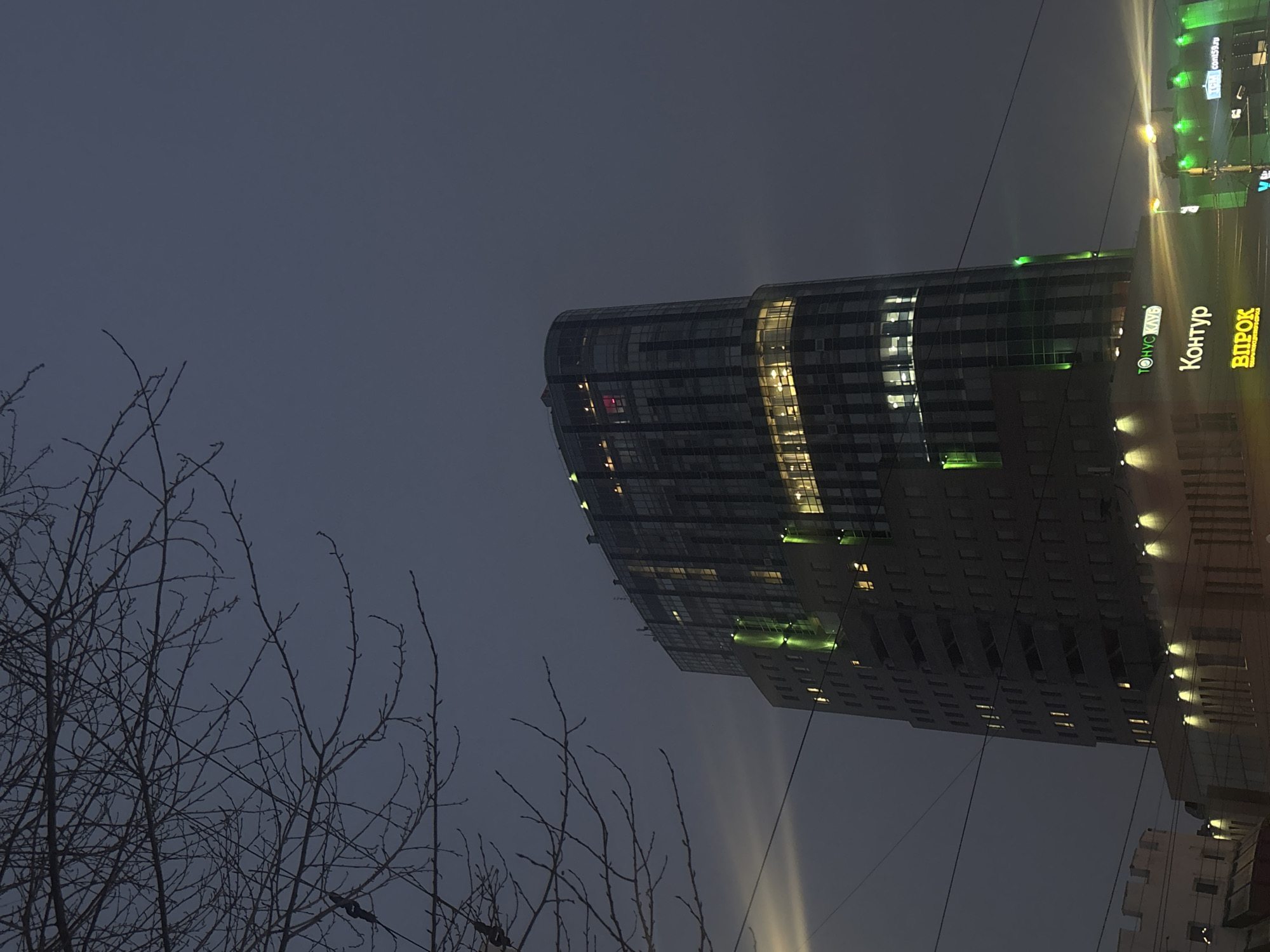 The image captures a nighttime view in Perm, Russia, featuring a tall, modern building and bare tree branches in the foreground. The building is illuminated with lights, with the sign "Контур" visible. The sky is dark and overcast. Electrical lines are also visible.