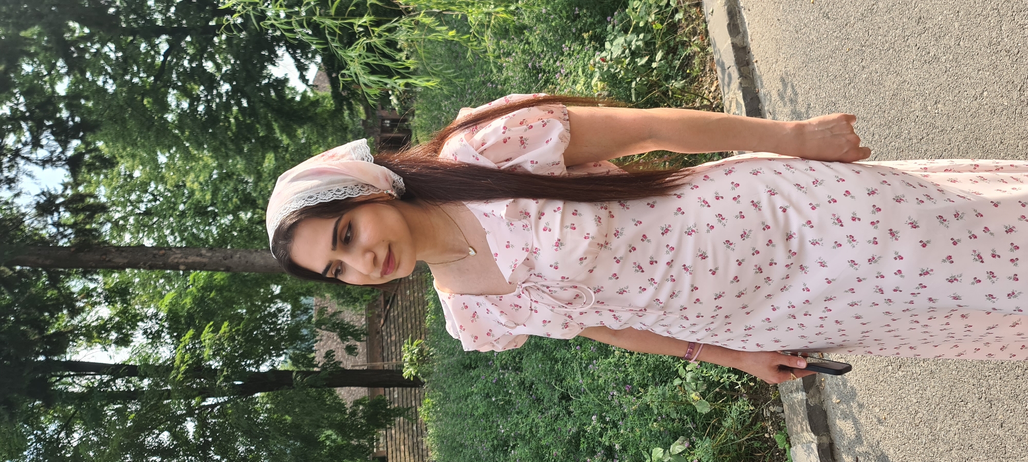 A young woman stands in an outdoor setting with a blurred background of trees and greenery. She is wearing a light pink, floral-patterned dress with puff sleeves and a lace-trimmed headscarf. A thin, heart-shaped necklace with a small gem is visible around her neck. In her right hand, she holds a dark-colored smartphone. She is looking to her left with a slight smile. The ground in front of her is a paved path transitioning to a rougher surface with small stones. The overall scene suggests a casual, natural environment, possibly a park or garden area within the city of Shaki, Azerbaijan, given the architectural hints in the background.