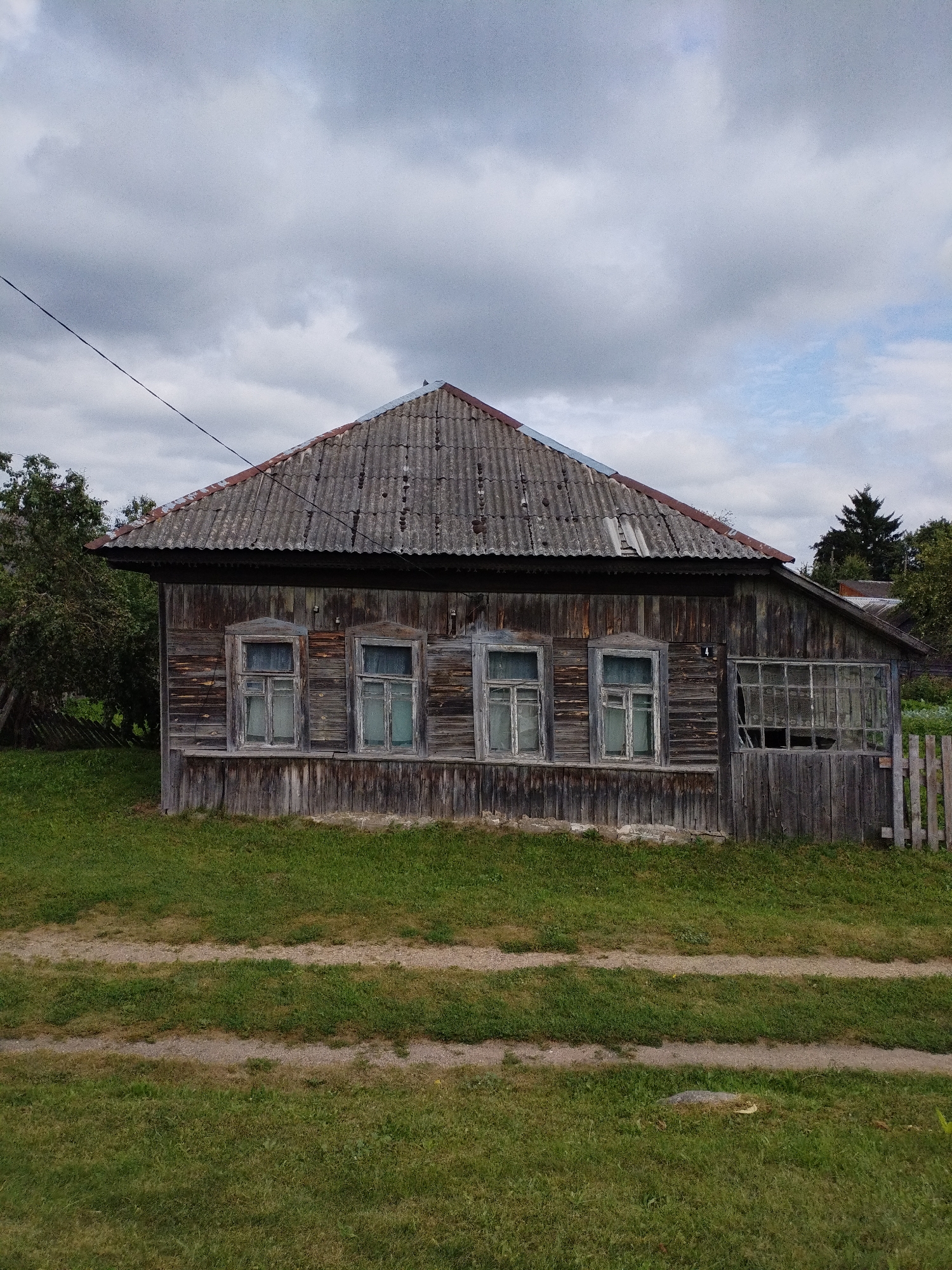 A weathered, wooden, single-story house with a corrugated roof sits in a grassy yard. The house has multiple windows, some of which are broken. A wooden fence runs along the right side of the property. The yard has a dirt path through it. The sky is overcast with clouds. The house is located in Bialynichy, Belarus.