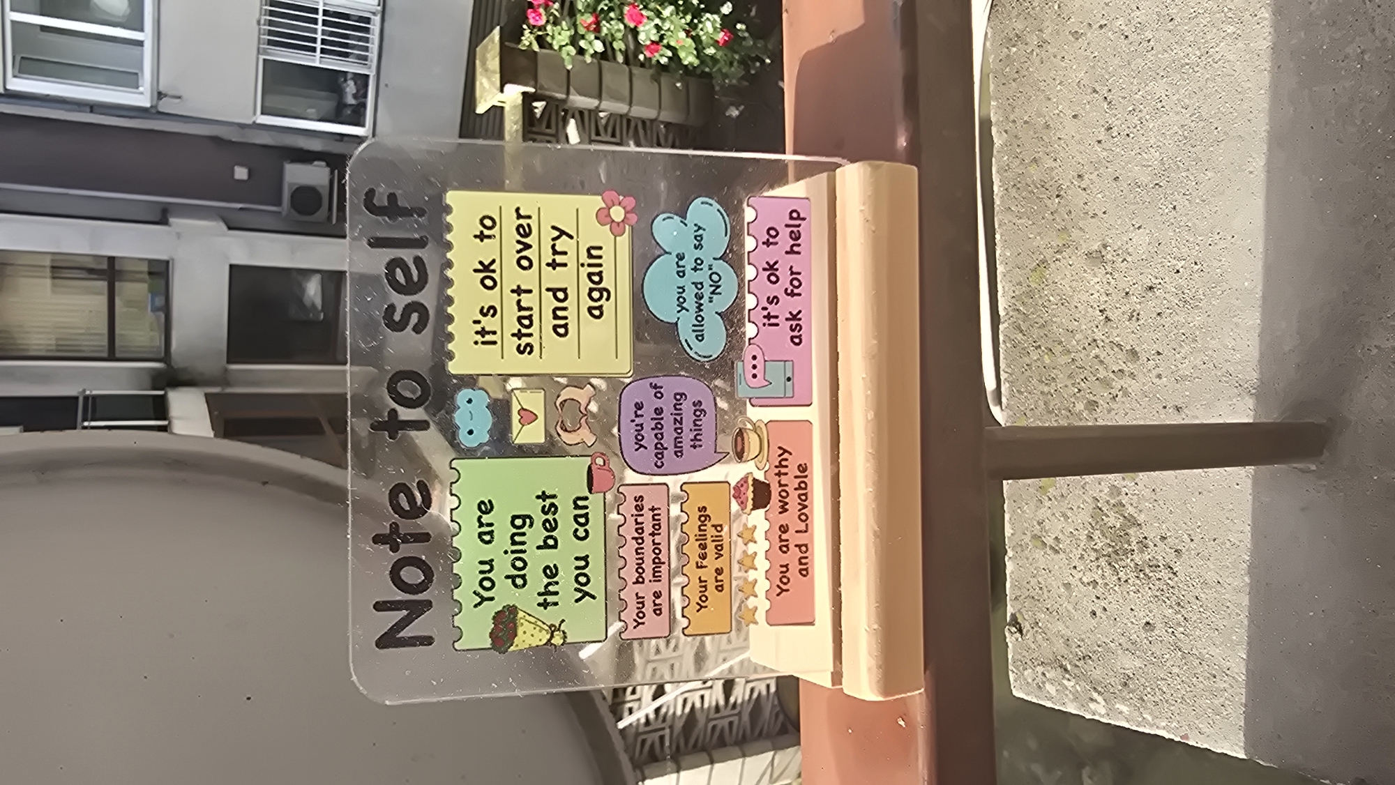 The image displays a clear acrylic sign with motivational messages, propped on a light wooden base, situated outdoors. The sign's main title, "Note to self," is written vertically down its left side.
In the background, blurred residential buildings with multiple windows and an air conditioning unit are visible, suggesting an urban environment. A dark planter with vibrant red and green flowers is also present. The foreground reveals a textured concrete surface, likely a balcony or window ledge, with distinct shadows indicating bright daylight.
The acrylic sign is decorated with various colorful, post-it note-style bubbles, each containing an uplifting phrase:
* A yellow bubble states: "it's ok to start over and try again".
* A light blue cloud-shaped bubble reads: "you are allowed to say 'NO'".
* A pink bubble advises: "it's ok to ask for help".
* A green bubble encourages: "You are doing the best you can".
* A purple bubble reminds: "you're capable of amazing things".
* A light brown bubble emphasizes: "Your boundaries are important".
* A light yellow bubble affirms: "Your feelings are valid", accompanied by four yellow stars.
* A light orange bubble, featuring a cupcake icon, declares: "You are worthy and Lovable".
Additional small illustrations, such as a blue cloud, a heart-envelope, a coffee mug, and a small pink flower, are scattered across the sign. The scene is well-lit, suggesting it is daytime with sunny weather.