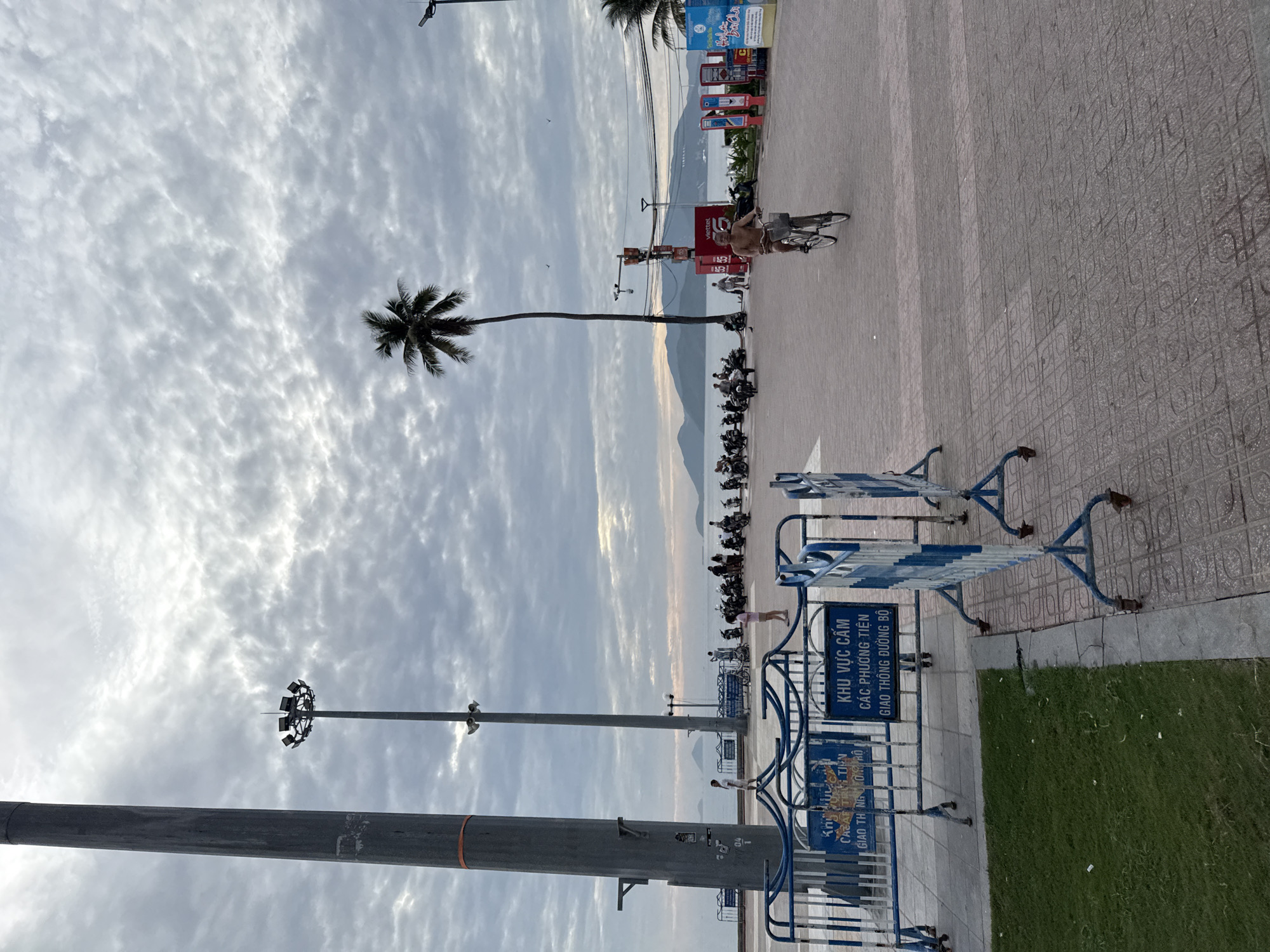 The image depicts a cloudy day scene along the coast of Nha Trang, Vietnam. In the foreground, a pathway is sectioned off by blue and white metal barriers, with a patch of green grass to the left. Beyond the barriers, a paved area features several people and parked motorcycles. A man is seen riding a bicycle towards the viewer, his upper body bare. A tall, slender palm tree stands prominently against the sky, which is filled with textured clouds. In the distance, the sea is visible with a hazy mountain range on the horizon. Two large light poles, likely for illuminating the area, are also visible. Signs with Vietnamese text are present, indicating restricted areas.