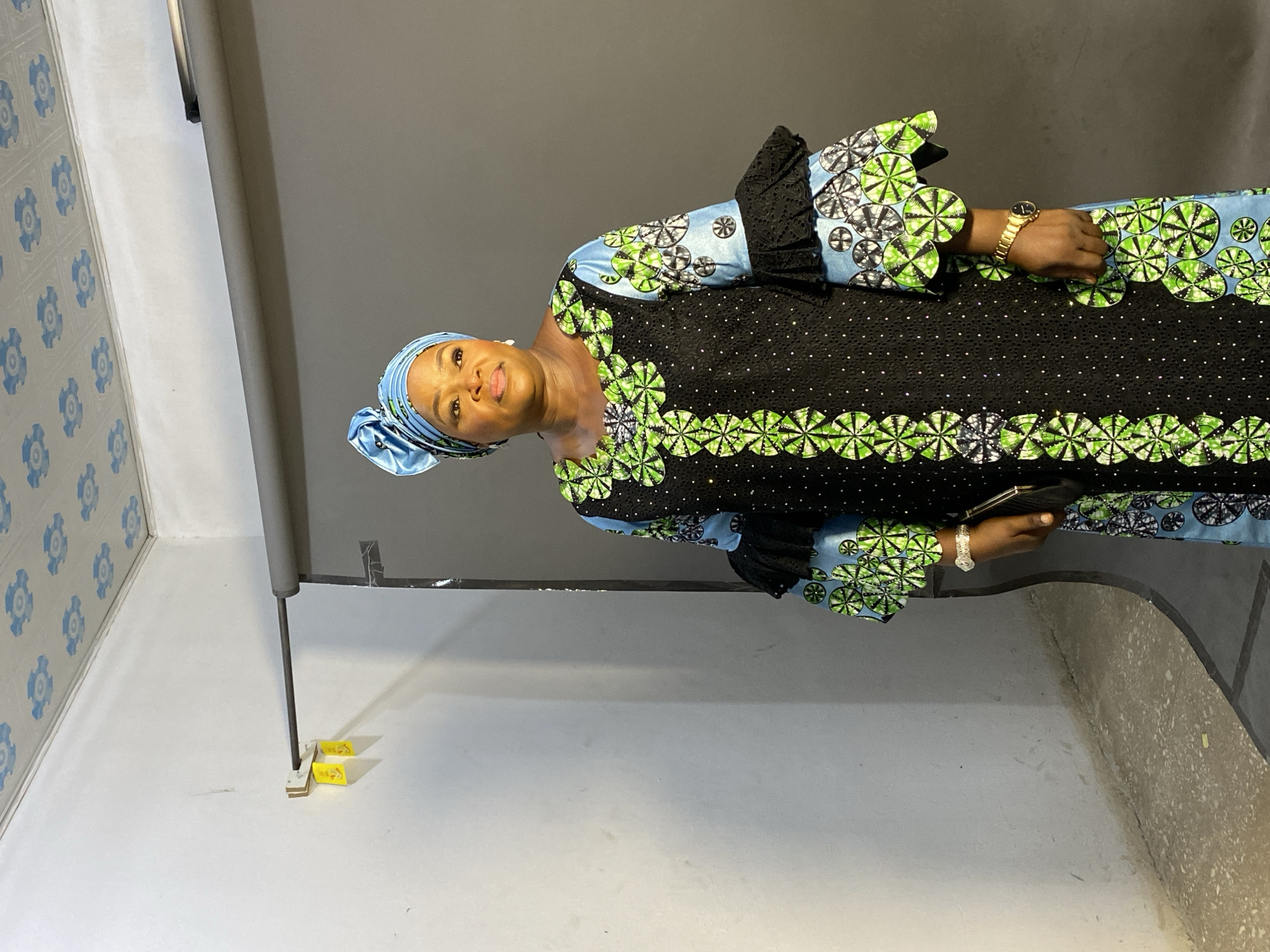 A middle-aged woman stands in a room, likely a studio, with a patterned blue head covering and a long, black dress with green and blue circular designs. The dress has a black lace trim around the sleeves and neckline. The woman is holding a black object in her right hand, possibly a phone or wallet. She is wearing a gold wristwatch on her left wrist and a silver bracelet on her right wrist.
The room has a light gray wall on the left with blue floral patterns. Behind the woman is a dark gray backdrop with a support stand visible on the lower left of the frame. The floor is primarily light gray, with a darker, textured area to the right of the backdrop.
The location is identified as Gura Top, Nigeria.