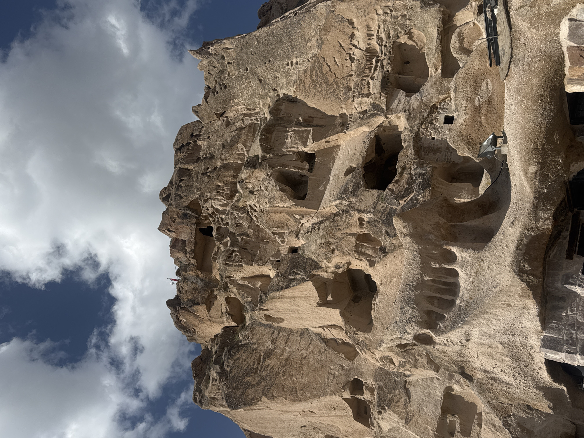 This image shows a rock formation with carved out spaces in Uçhisar Beldesi, Turkey, likely a historic cave dwelling site. The rock face is textured and weathered with numerous holes and rooms. The sky is visible on the left side with a mix of blue and white clouds. A structure with dark metal poles is attached to the side of the rock formation.