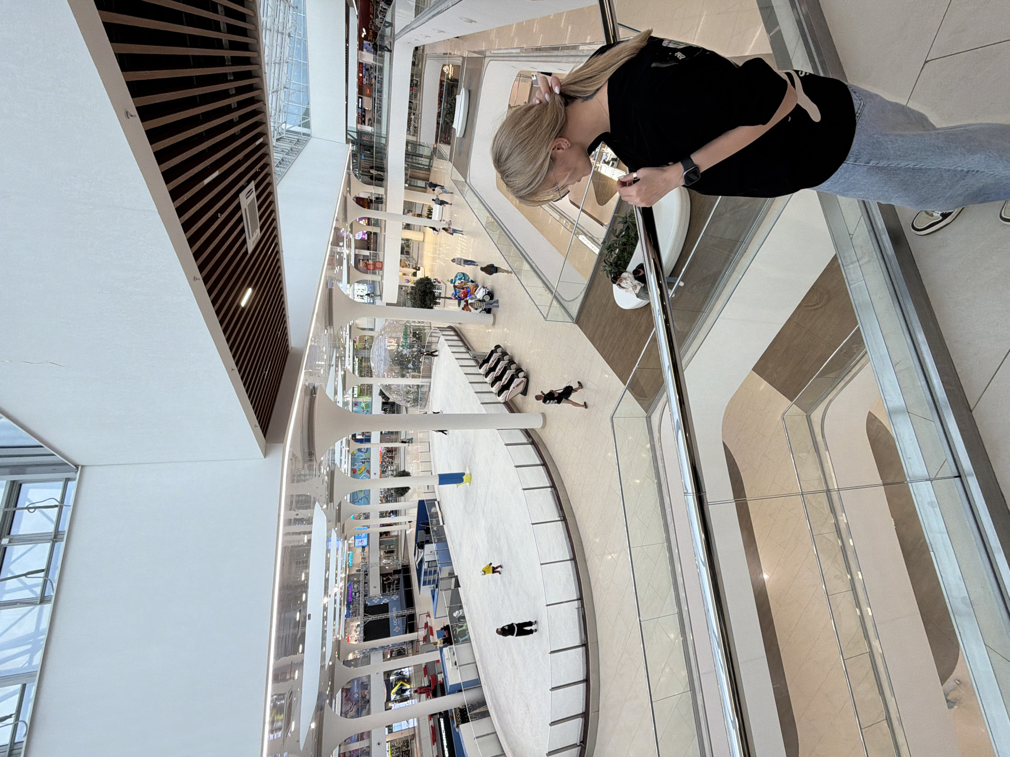 The image depicts an indoor scene within a multi-level shopping mall, likely Nizhny Novgorod, Russia. The perspective is from an upper level, looking down and across. The main subject is a young woman with blonde hair tied in a ponytail, wearing a black t-shirt, blue jeans, and white sneakers. She is leaning over a railing, looking downwards. Her hair is styled in a ponytail that falls over her shoulder. She is wearing glasses and a smartwatch on her left wrist.
The mall features several levels with various shops visible in the background. There are escalators and walkways connecting the different floors. A prominent feature on one of the lower levels is an ice rink, where several individuals are visible, some of whom appear to be skating or have fallen. One person in a yellow shirt is lying on the ice, and another in a black and white outfit is also on the ice. Further in the background, other shoppers are seen walking around.
The setting is brightly lit, suggesting it is daytime. The architecture of the mall includes large windows, modern lighting fixtures, and decorative elements such as large circular pillars and wood-paneled ceilings. Reflections are visible on some of the surfaces, indicating the presence of glass and polished materials. No specific time of day or weather conditions are discernible from the indoor environment. The general mood seems to be one of casual activity and shopping. No discernible text is clearly legible within the image.