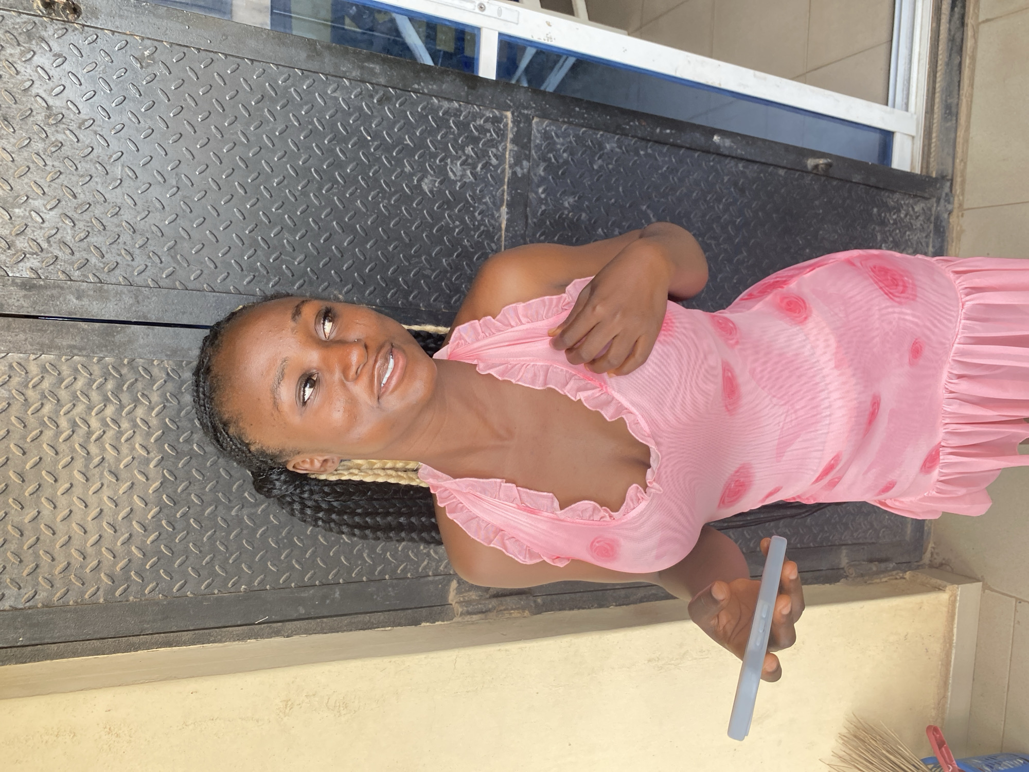 A young Black woman is depicted lying on a dark, textured metal surface, with the image rotated 90 degrees counter-clockwise. She is smiling broadly and gazing directly at the camera. Her hair is styled in long, dark braids, interspersed with sections of blonde or light brown braids. She is wearing a light pink, semi-sheer dress with ruffled detailing along the neckline and shoulders, and a faint pattern of darker pink rosettes or polka dots across the fabric.
In her right hand, she holds a light blue or grey smartphone. The background behind her consists of more of the dark, diamond-plate-like metal surface. Above her head, a window or glass panel with a white frame is visible, suggesting an indoor or sheltered outdoor setting. In the bottom right corner of the frame, a cluster of light-colored, possibly straw, brushes or brooms is partially visible. The lighting indicates it is daytime. The woman's expression conveys happiness and ease. The scene is located in Jos, Nigeria. There is no visible text in the image.