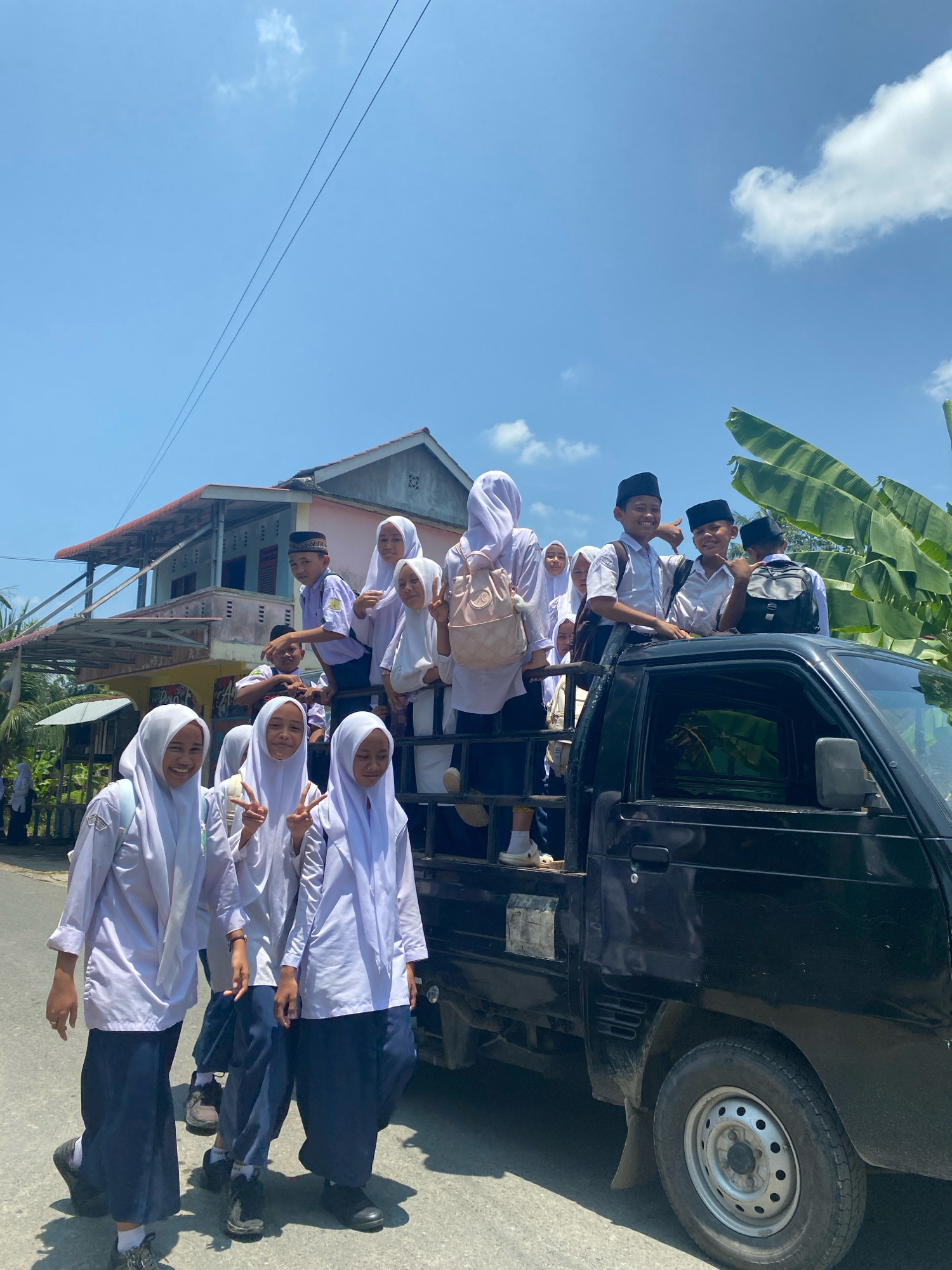 In Karang Gading, Indonesia, a group of Indonesian schoolchildren, predominantly female and wearing white hijabs and uniforms, are standing in the back of a black pickup truck. Some of the children are smiling and making peace signs. The truck is parked on a paved road, with a building and lush greenery visible in the background. The sky is clear blue with a few white clouds.