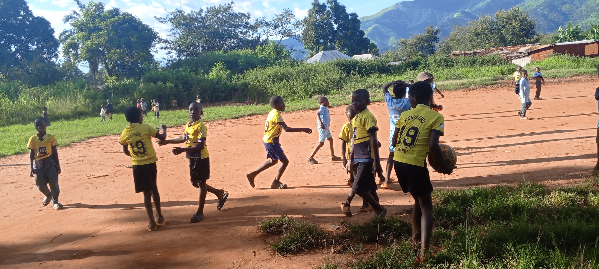 The image depicts an outdoor scene in Ngalula, Democratic Republic of the Congo, featuring a group of approximately 15-20 children and young adolescents on a reddish-brown dirt field. Several individuals wear yellow and black soccer jerseys, some bearing "DORTMUND" text and numbers (e.g., "19 BRANDT," "9," "1&1"). Others wear casual attire, including light blue shirts and shorts. One individual in the foreground, facing away from the camera, holds a deflated or worn soccer ball.
The children are in various states of movement, suggesting active play or gathering. Some are walking, others appear to be running or gesturing. Shadows are cast on the dirt ground from the individuals and implied overhead sun.
The environment includes a strip of green grass and dense green foliage (bushes and trees) bordering the dirt area. In the mid-ground, simple structures with corrugated metal roofs and some white tent-like structures are visible among the vegetation. The background features prominent green mountains under a sky with scattered white clouds, indicating a clear day.