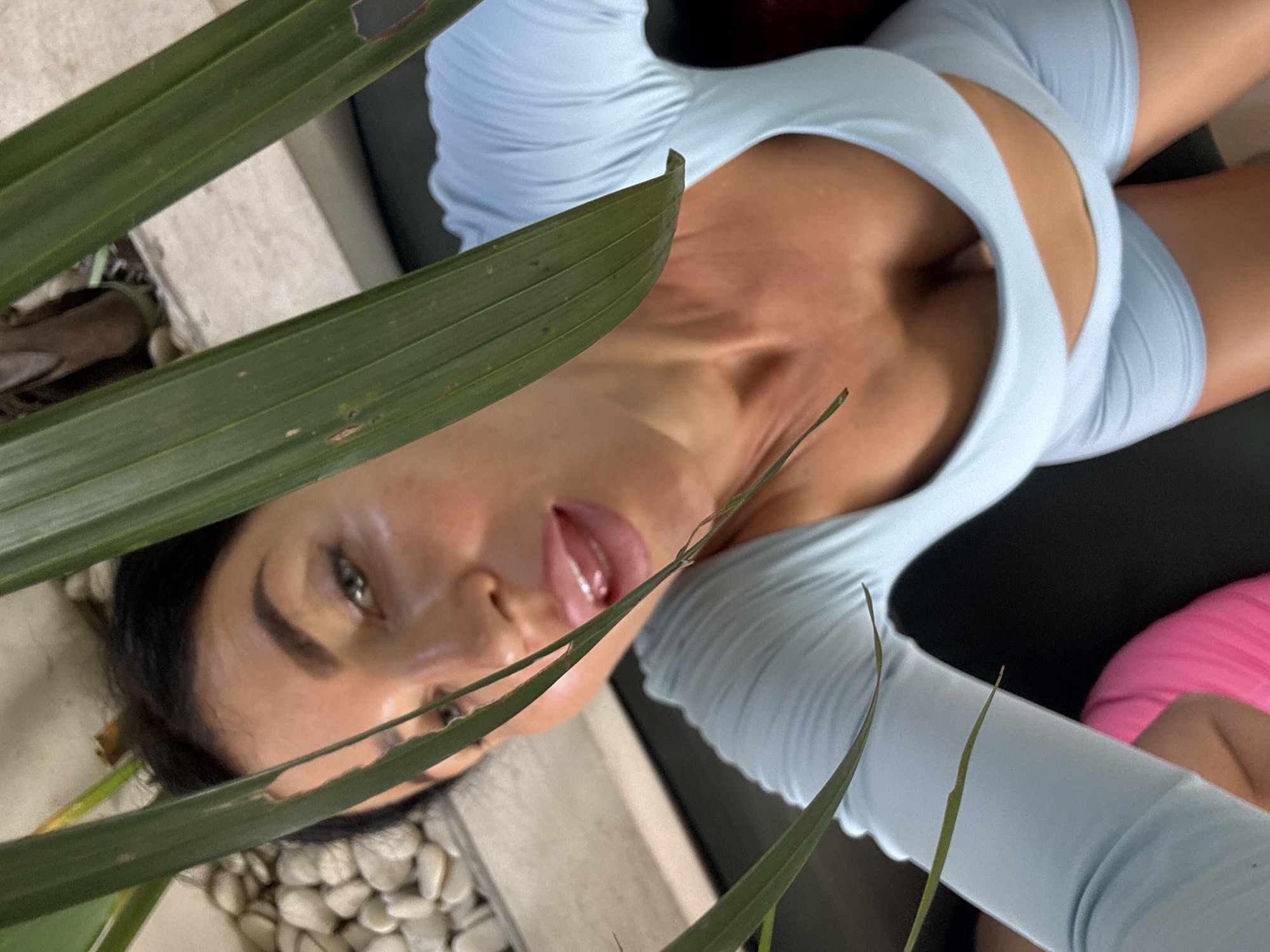 A woman with dark hair and green eyes, wearing a light blue long-sleeved crop top and matching shorts, poses for a selfie in an outdoor setting. She is partially obscured by large green leaves. The background shows a textured surface with white stones and a hint of brown wood. The image appears to have been taken in Mengwi, Indonesia, likely in a garden or courtyard area.