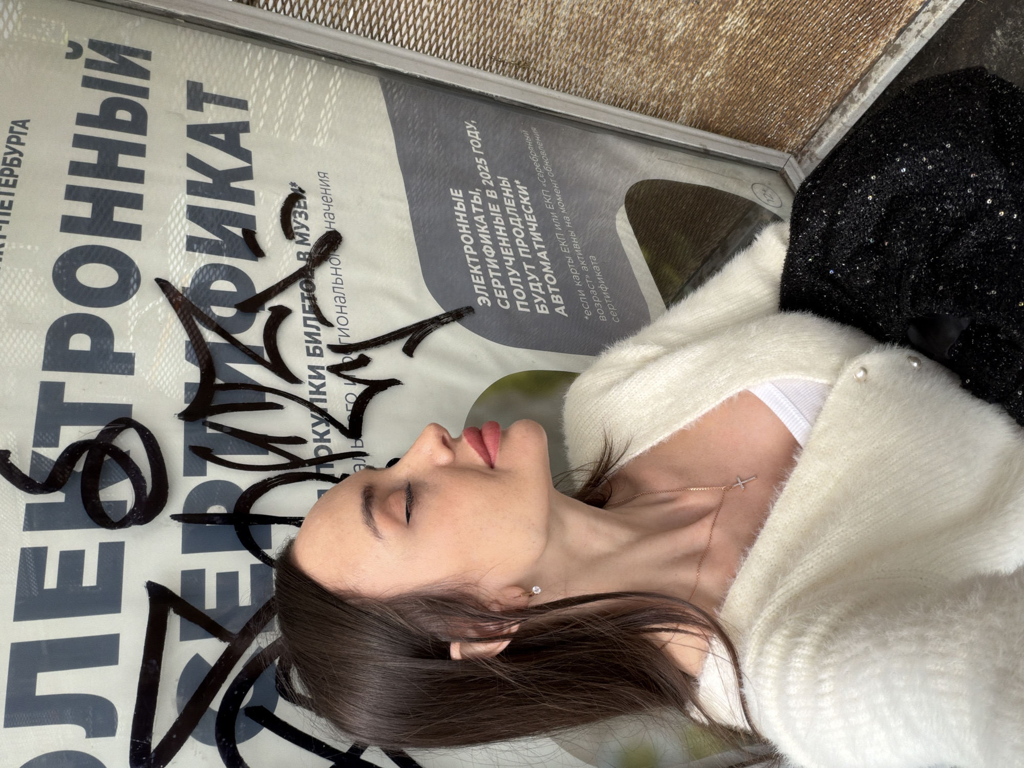 The image shows a woman lying next to a poster in Saint Petersburg, Russia. The poster advertises electronic certificates for museum tickets. The woman is wearing a white sweater, a white tank top, and a necklace. She is lying on her back with her eyes closed.