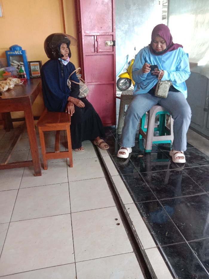 Two people are seated on stools. On the left, an older person wears a helmet, dark blue clothing, and sandals. A purse hangs from her shoulder. To the right, a younger person in a light blue sweatshirt, jeans, and white sandals is using a mobile phone. A purple hijab covers her head. A yellow motorcycle is visible in the background. A brown wooden table and maroon cabinet also appear. The floor is tiled with light and dark tiles, separated by a metallic strip.