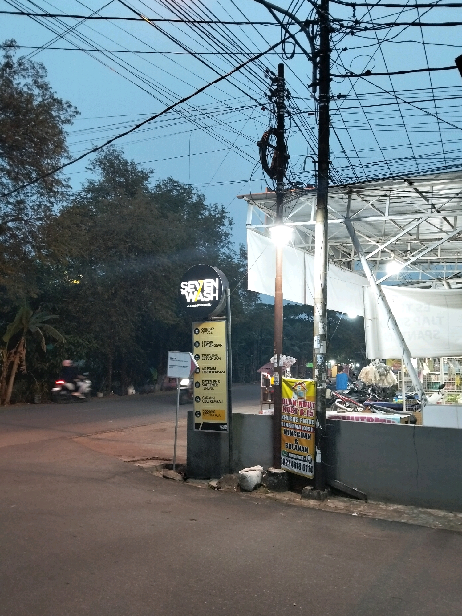 The image depicts a street scene at dusk in Pontianak, Indonesia. A prominent sign for "SEVEN WASH - LAUNDRY EXPRESS" is visible, along with smaller signs detailing services like "ONE DAY SERVICE," "TERPANTAU CCTV 24 JAM," "AIR PDAM TERFILTERISASI," "DETERJEN & SOFTEENER TERBAIK," and "GARANSI CUCI KEMBALI." Another vertical banner advertises "OLAN NDUT KOST B.15 KHUSUS PUTRA MENERIMA KOST MINGGUAN & BULANAN" with a contact number. A motorcycle is being ridden on the road, and there are other parked motorcycles and a small market stall in the background. Overhead, a complex network of electrical wires crisscrosses the sky, supported by utility poles. The overall environment suggests a commercial area with residential housing nearby.