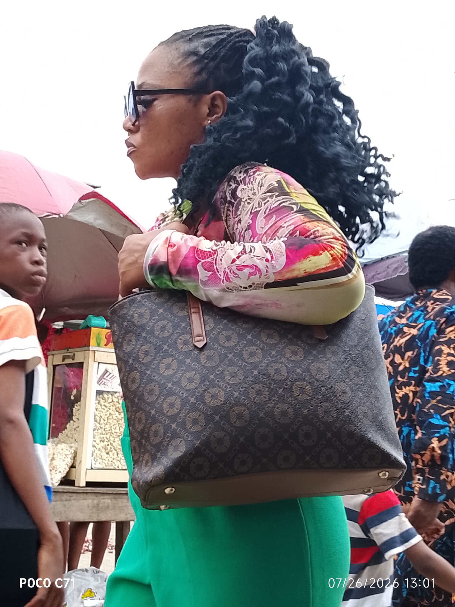 In Lekki, Nigeria, a woman with dark, braided hair and large sunglasses stands in profile, holding a large, patterned brown handbag with a light brown trim and shoulder strap. She wears a colorful, long-sleeved shirt with a pink, white, and green abstract print. Her lower body is obscured by a bright green skirt. To her left, a young boy with short black hair looks towards the viewer, wearing a striped shirt with white, orange, and dark stripes. Behind the woman and boy, a stall displays various snacks, possibly nuts, in clear plastic bags. The background is a light, overcast sky. Another person wearing a blue and orange patterned garment is visible to the right. The scene appears to be an outdoor market.