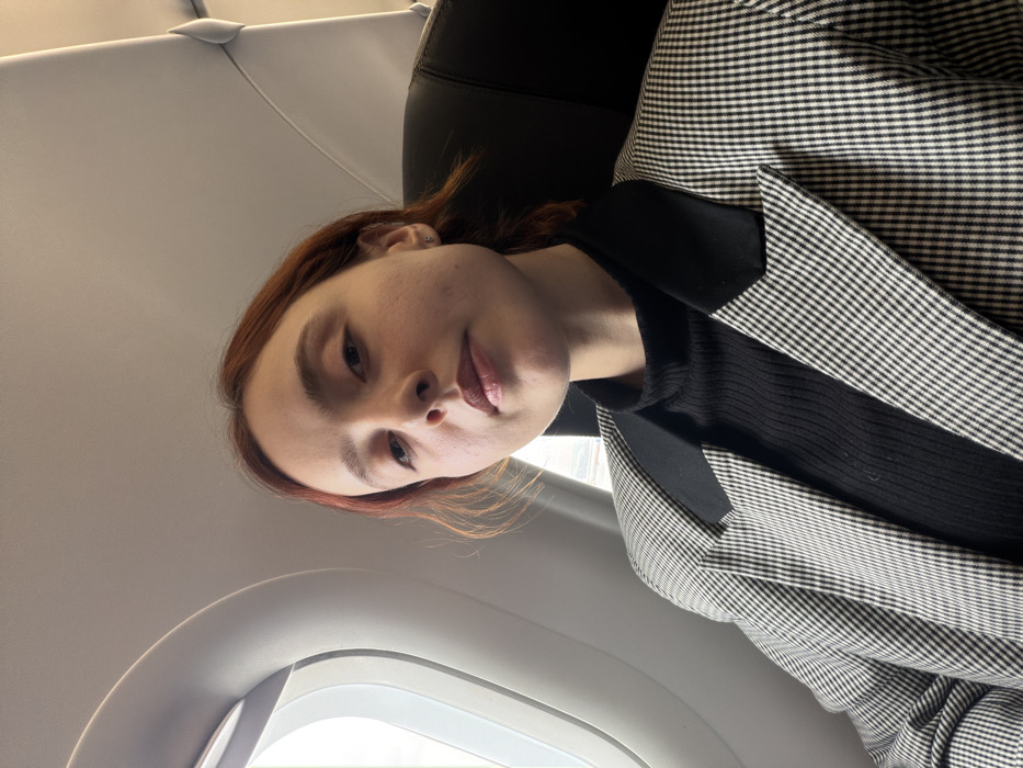 A female individual is depicted in a close-up shot, seated within the cabin of an aircraft. The location is on board an aircraft, likely at or near Sabiha Gökçen International Airport (SAW), which serves the Tuzla district of Istanbul, Turkey.
The subject is a young adult female with fair skin and reddish-brown hair, styled away from her face. She wears a small stud earring in her left earlobe. Her lips are lightly colored, and her gaze is directed towards the camera with a neutral expression.
She is dressed in a black ribbed turtleneck sweater, overlaid by a black and white small-checkered blazer with solid black lapels.
To her left, an oval-shaped airplane window is visible, revealing an overexposed external scene indicating daylight. The interior structure of the aircraft, including a portion of the ceiling and an overhead panel, is discernible. A dark-colored aircraft seatback is partially visible to her right. The image is captured from a slightly low angle, with the subject occupying the majority of the frame.