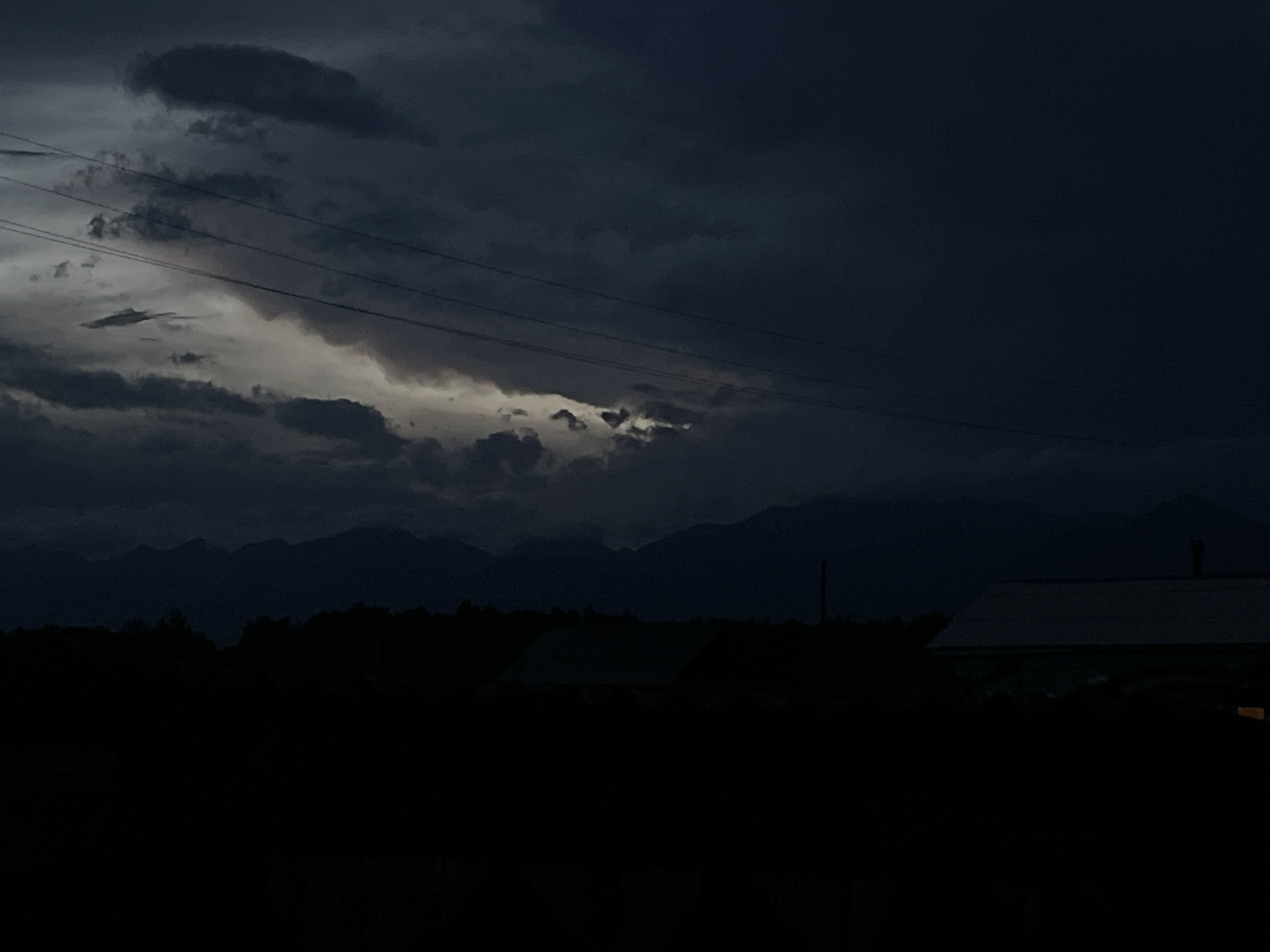 The image depicts a dark, cloudy sky over a silhouetted landscape of mountains, trees, and buildings. The sky is filled with ominous, dark clouds, with a sliver of lighter, illuminated cloud peeking through in the upper center of the frame. Several power lines stretch horizontally across the sky. Below the sky, a dark silhouette of a treeline is visible, separating the sky from the foreground. In the foreground, the dark silhouettes of several buildings are present, one with a visible roofline. The overall impression is one of impending weather or twilight. The location is described as Unknown, Unknown.