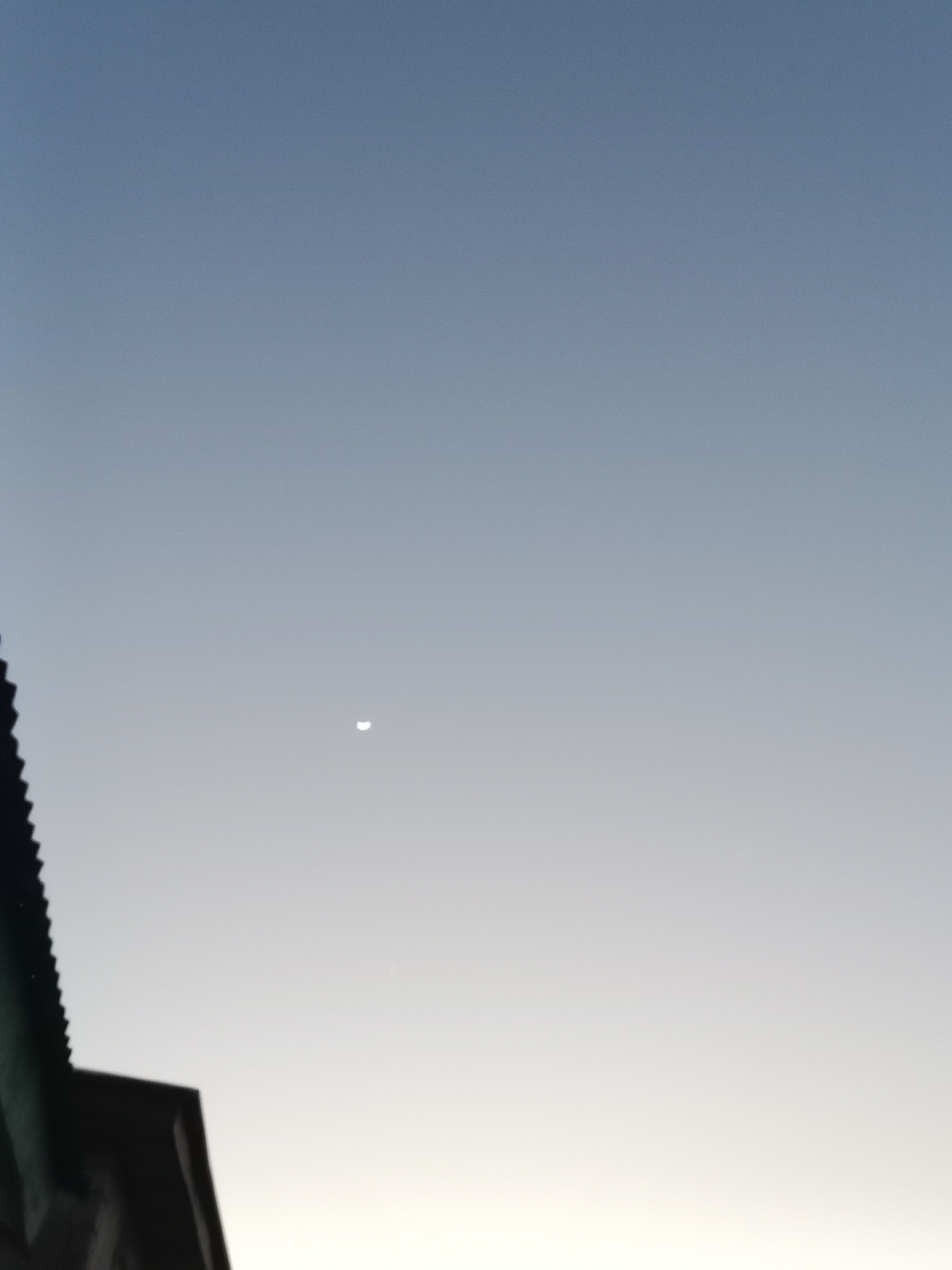 The image shows a crescent moon in a clear sky, appearing as a thin sliver of light against a gradient of blue to white. The sky suggests either dawn or dusk. In the foreground, a portion of a building with a dark, sloped roof is visible on the left side of the frame, along with the upper part of a dark structure that appears to be another building or roof. The overall scene is simple, focusing on the celestial body and the transition of light in the sky, with man-made structures providing a grounding element in the lower left corner. The location is specified as Lokoja, Nigeria. There is no visible text in the image.