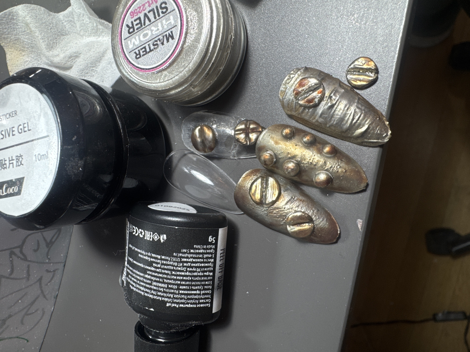 The image displays a close-up, top-down view of nail art supplies and decorated nail tips laid out on a dark grey surface, likely a desk or workbench.
In the upper left, a black cylindrical bottle is partially visible, labeled "STICKER ADHESIVE GEL" and "10ml." Next to it, a round, silver-colored container with a lid is prominently featured, bearing the text "MASTER HROM SILVER" and "art.2256," indicating a chrome or metallic nail powder. Below these, another black bottle, partially cropped, is visible. Its label, largely in Russian, identifies it as "PEEL OFF BASE" and specifies "5g" volume, "Made in China," and lists ingredients such as Acrylates Copolymer and Cellulose Acetate Butyrate. Further text includes application instructions ("apply a thin layer to nails without coating," "dry with a lamp for 60 seconds"), warnings ("for external use only," "keep away from children," "keep away from UV rays"), and manufacturer details: "Произведено для: ИП Фёдорова Евгения Борисовна," with an address in Moscow (121357, Россия, Москва, пр-кт Кутузовский, 20 кр 1), email "imennails@mail.ru," and a "Срок годности: 5 лет" (shelf life: 5 years).
The main subjects are several artificial nail tips, some transparent and others elaborately decorated. The decorated tips exhibit a metallic, steampunk, or industrial design, appearing aged or tarnished in shades of silver, bronze, and oxidized gold. They are embellished with textured surfaces and various screw-head or rivet-like ornaments. One small, round, metallic screw-head ornament is also seen loose on the grey surface.
A crumpled white tissue or wipe is visible in the top left corner of the image. The lighting appears to be artificial, casting soft shadows that highlight the textures and metallic sheens of the nail art. The scene suggests a creative activity involving the preparation and display of intricate nail designs.