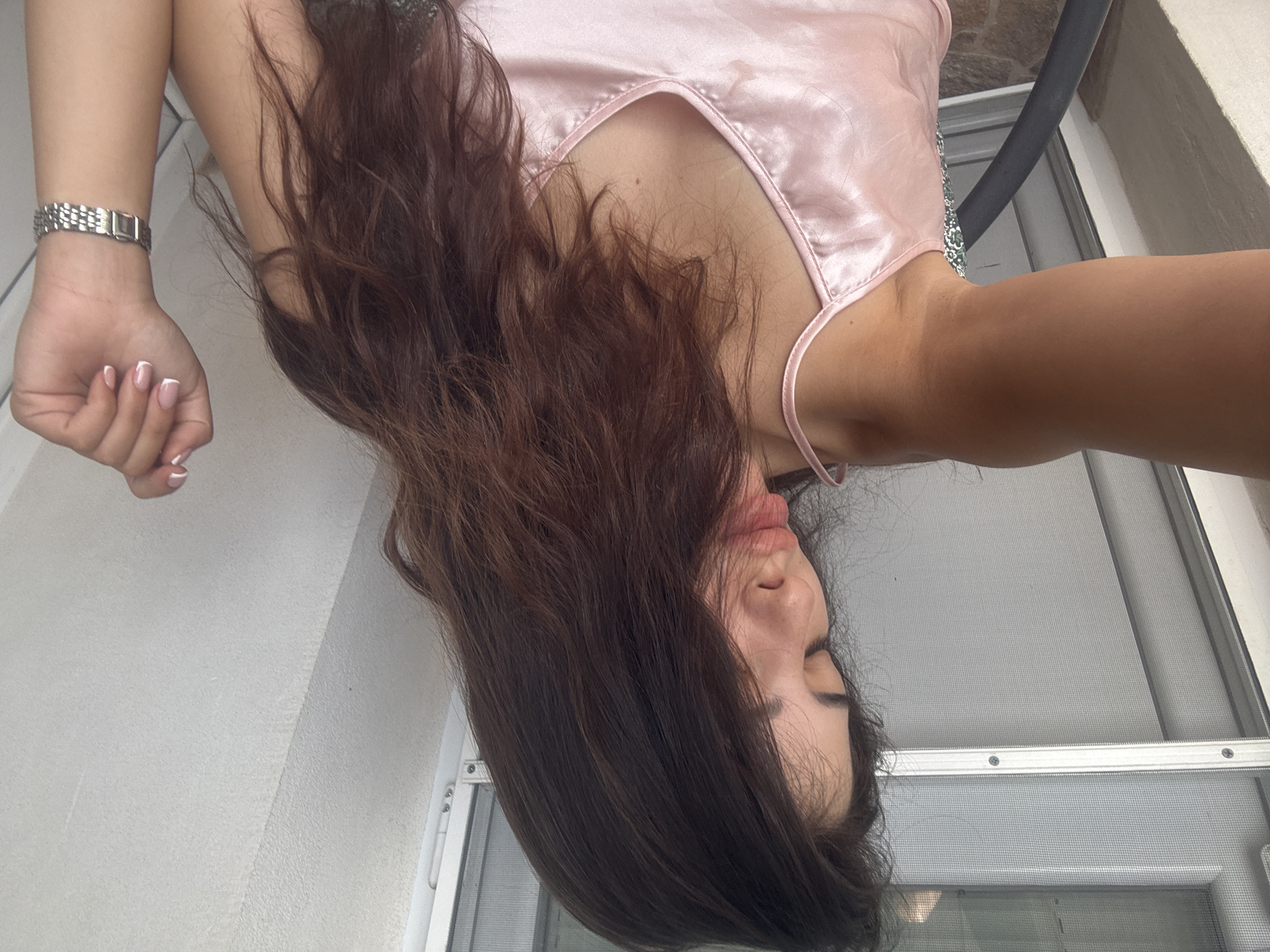 A person is lying down, possibly outdoors on a patio or balcony, in Plovdiv, Bulgaria. The person has long, dark brown hair that cascades around their face and shoulders. They are wearing a light pink, silky camisole that appears to be pajamas or loungewear. The camera angle is from above, looking down at the person.
Visible details include the person's face, with their eyes closed and lips slightly parted, suggesting a relaxed or possibly sleepy state. They are wearing a silver watch on their left wrist and their fingernails are painted with a French manicure. Behind the person, there is a gray screen door and a white wall, indicating an outdoor or semi-outdoor setting. The lighting suggests it is daytime, but the specific time of day and weather conditions are not clearly discernible. No text is visible in the image. The scene is casual and intimate, focusing on the person's personal space and relaxed posture.