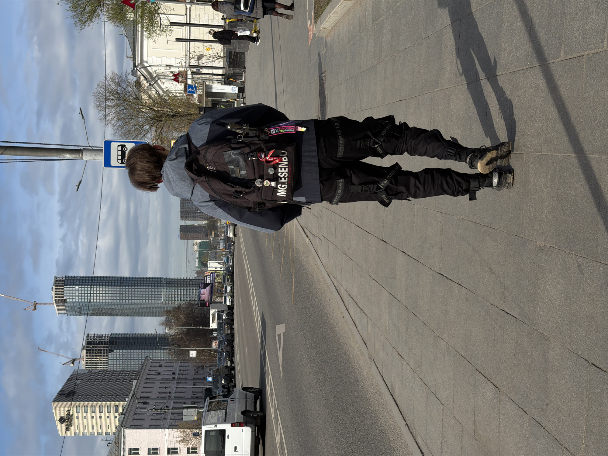 A person is walking on a sidewalk next to a road in what appears to be an urban environment. The person is wearing dark clothing and a backpack, and their shadow is cast on the sidewalk. A bus stop sign is visible nearby. In the background, there are tall buildings, including ones under construction with cranes. The road has white lane markings and several vehicles. The location is likely Khimki, Russia, based on the background and available context.