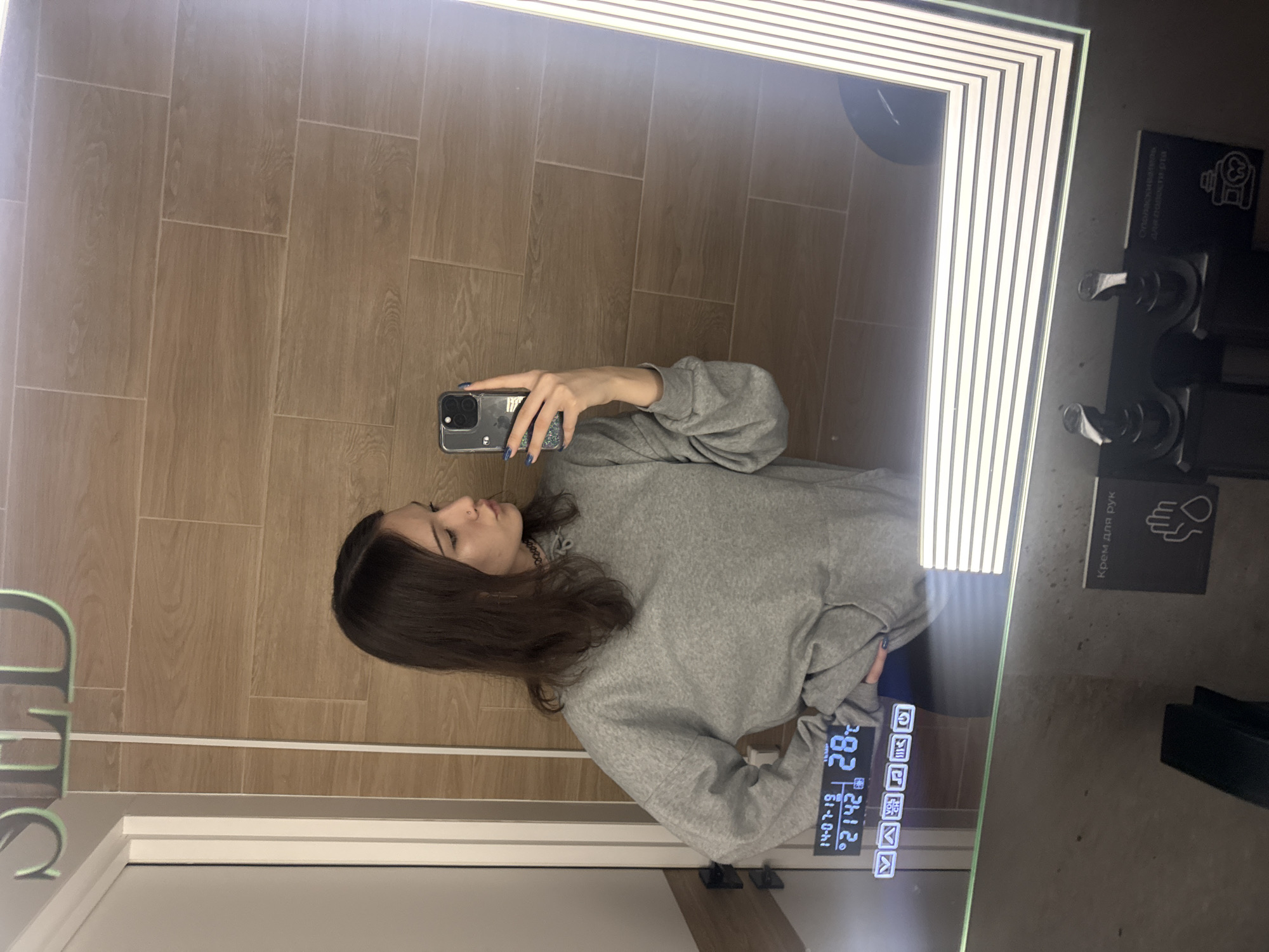 The image displays a mirror selfie taken by a young woman in what appears to be a modern bathroom or changing room. The woman, with dark brown hair, is wearing a gray long-sleeved sweatshirt and has blue nail polish on her fingers. She holds a dark-colored smartphone with a clear, sparkly case to capture her reflection. Her face is seen in profile, with a neutral expression as she looks towards the phone screen.
The background behind her reflection consists of light brown, wood-grain textured tiles arranged in a vertical pattern. The mirror itself is a 'smart' mirror, bordered by bright LED strip lights. On the lower right side of the mirror's surface, a digital display shows the date "14-07-19" (July 14, 2019), the time "21:42" (9:42 PM), and a temperature of "28°C". Below these, several touch-sensitive icons are visible, including those for volume control and music playback (play/pause, skip forward, skip backward). On the far left, the word "LED" is reflected in green letters.
To the right of the mirror, on an adjacent dark panel or frame, there are instructions and icons written in Cyrillic. One section clearly states "Крем для рук" (Cream for hands) with an icon of a hand and a drop, suggesting a hand cream dispenser. Above it, text reads "Специальное отделение для понести рта" (Special compartment for oral cavity/mouth), accompanied by an icon resembling an oral hygiene product holder. The setting appears contemporary and well-lit, consistent with a facility in Saint Petersburg, Russia, as indicated by the Cyrillic text.