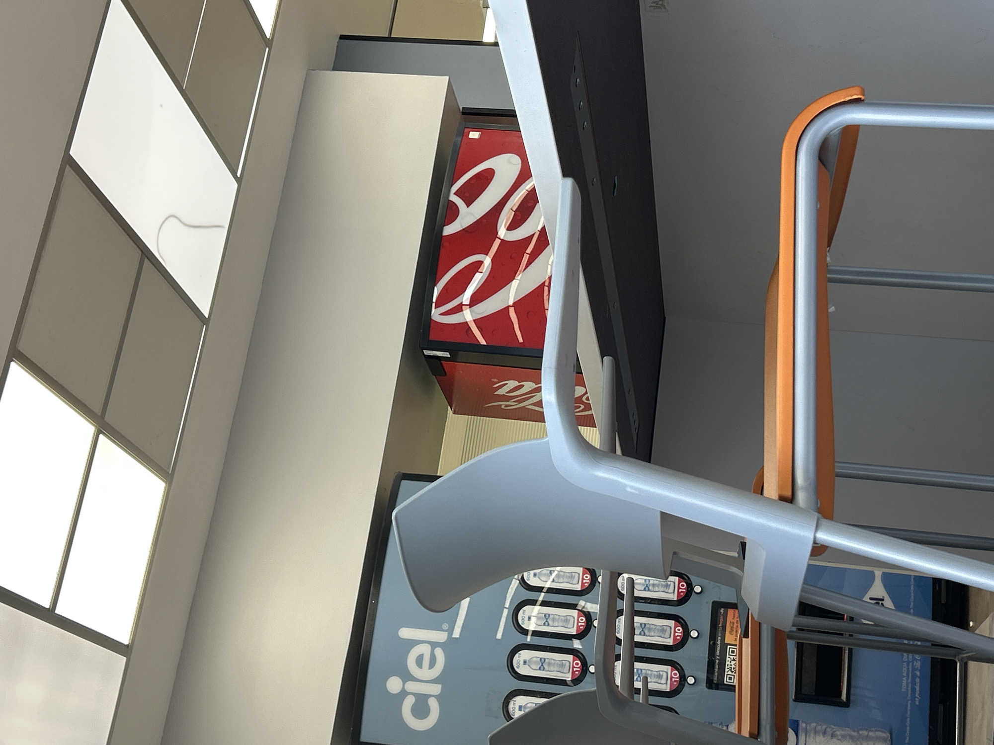The image shows the interior of a building with an angled geometric window design in the upper left. Next to the wall, there is a tall white column-like structure. Adjacent to it, there is a Coca-Cola branded refrigerator. Below the Coca-Cola fridge, there is a cooler branded "Ciel" with beverage options marked at "$10". In the foreground, there are a grey plastic chair and an orange-framed chair stacked together. The image is rotated at an angle.
