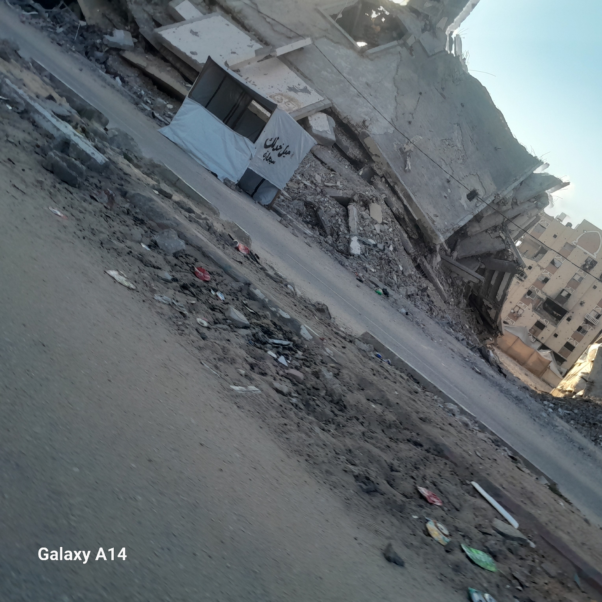 The image depicts the aftermath of destruction in the city of Khan Yunis, Palestinian Territories. A partially collapsed building, characterized by exposed concrete and rebar, dominates the right side of the frame. Debris litters the foreground and a road that runs diagonally across the scene. A white tarp with Arabic writing, possibly advertising or informational, is visible near the base of the damaged structure. The sky above is clear and blue. The overall scene conveys devastation and ruin, suggesting a recent destructive event. The "Galaxy A14" watermark is present in the lower left corner.