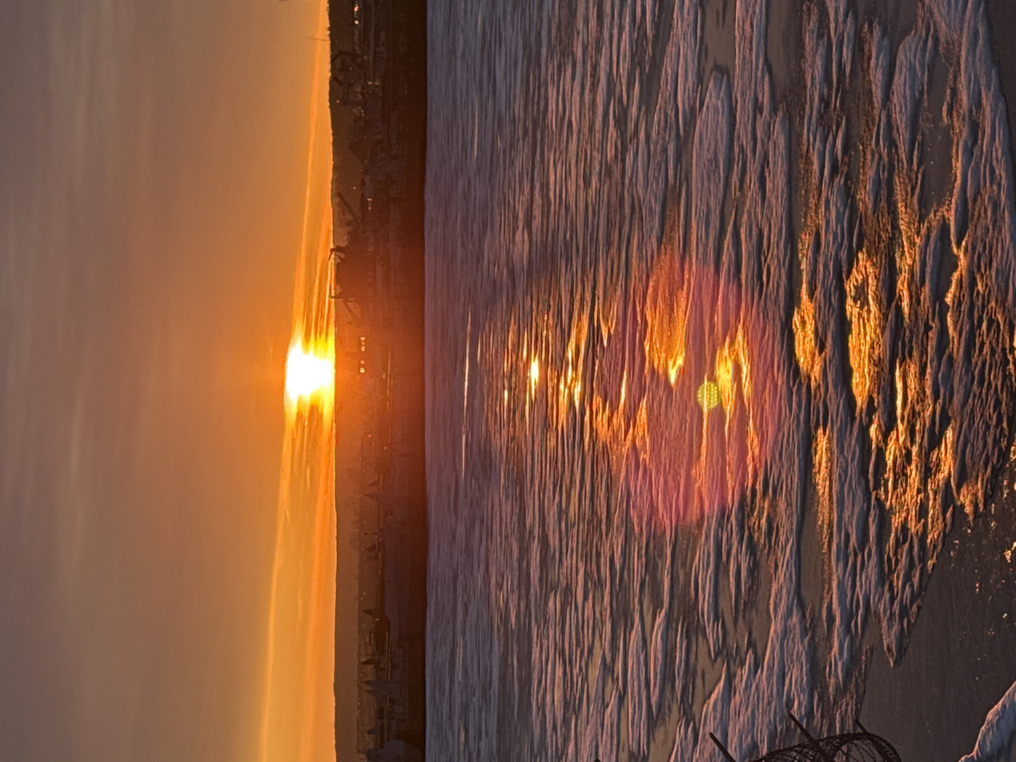 The image depicts a sunset over the Kama River in Chaikovsky, Russia. The sun is setting on the left side of the frame, casting a bright orange glow over the city skyline. The reflection of the sunset is visible on the surface of the Kama River on the right side. The water is patterned. In the bottom corner, there is razor wire.