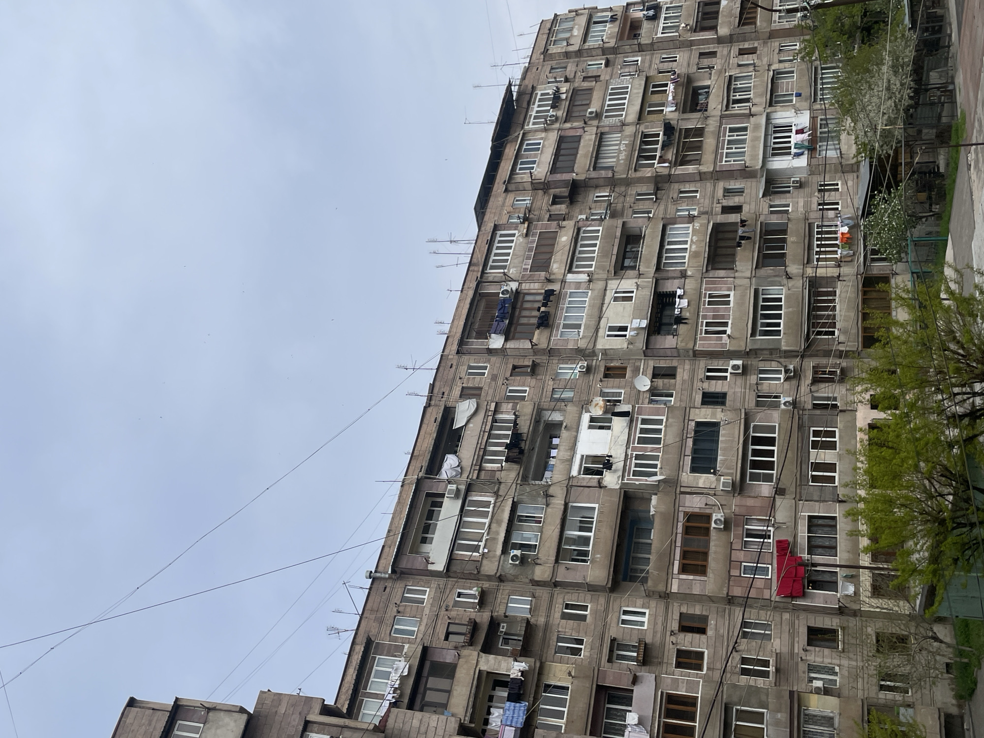 The image depicts a high-rise apartment building in Yerevan, Armenia. The building is characterized by its repetitive window patterns, each unit often featuring unique modifications like different window styles, satellite dishes, and air conditioning units. Laundry is visible hanging from some windows. The facade appears weathered and aged. The sky is overcast. Trees are visible on the right side of the frame, suggesting the building is situated near a street or park. Power lines cut across the sky in the lower left of the frame.