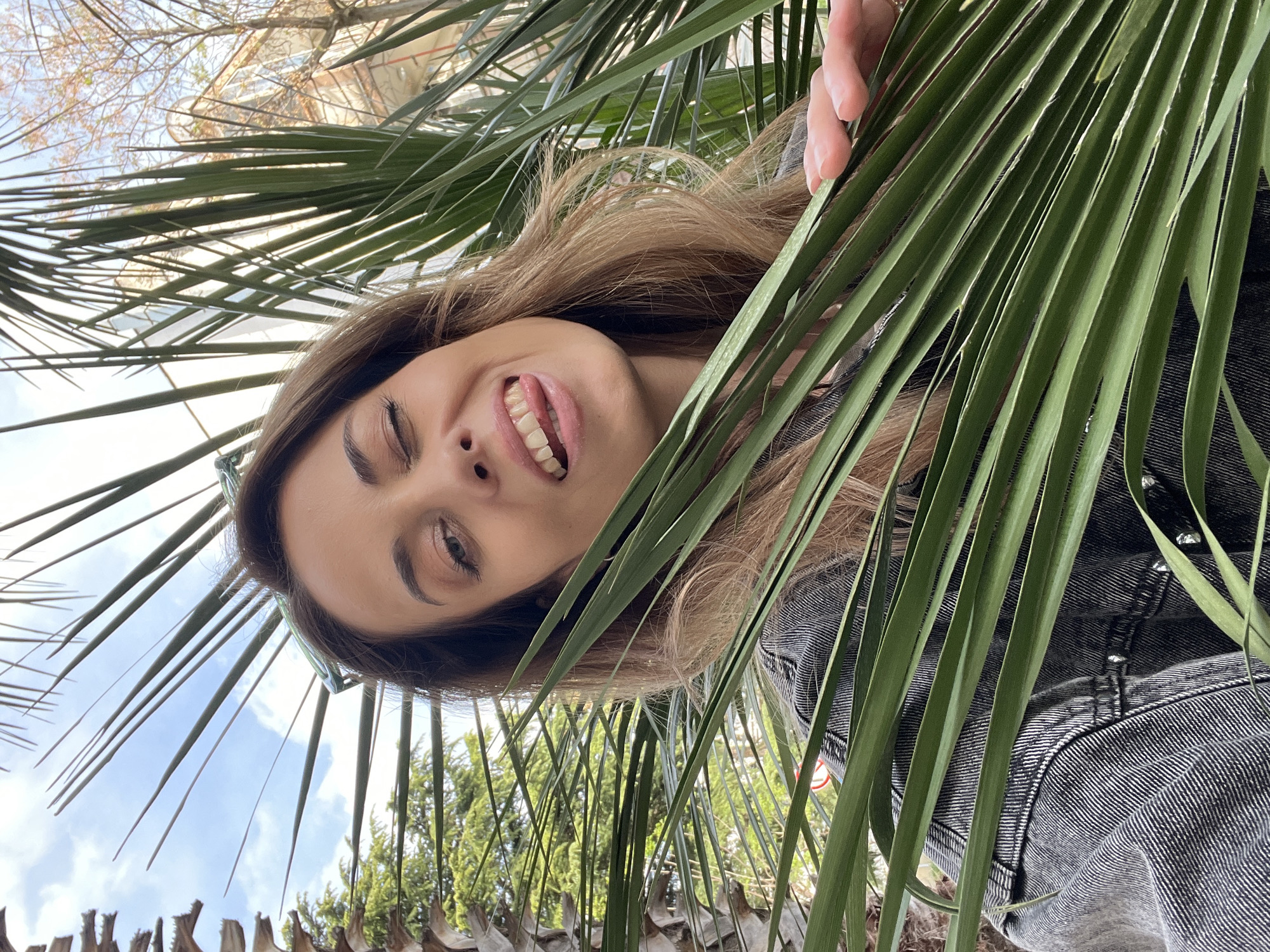 A photograph taken in Bar, Montenegro, depicts a young woman smiling with her tongue slightly protruding between palm leaves. She has brown hair, light skin, and wears a denim jacket. The sky is visible in the background. A palm tree is framing the bottom of the image.