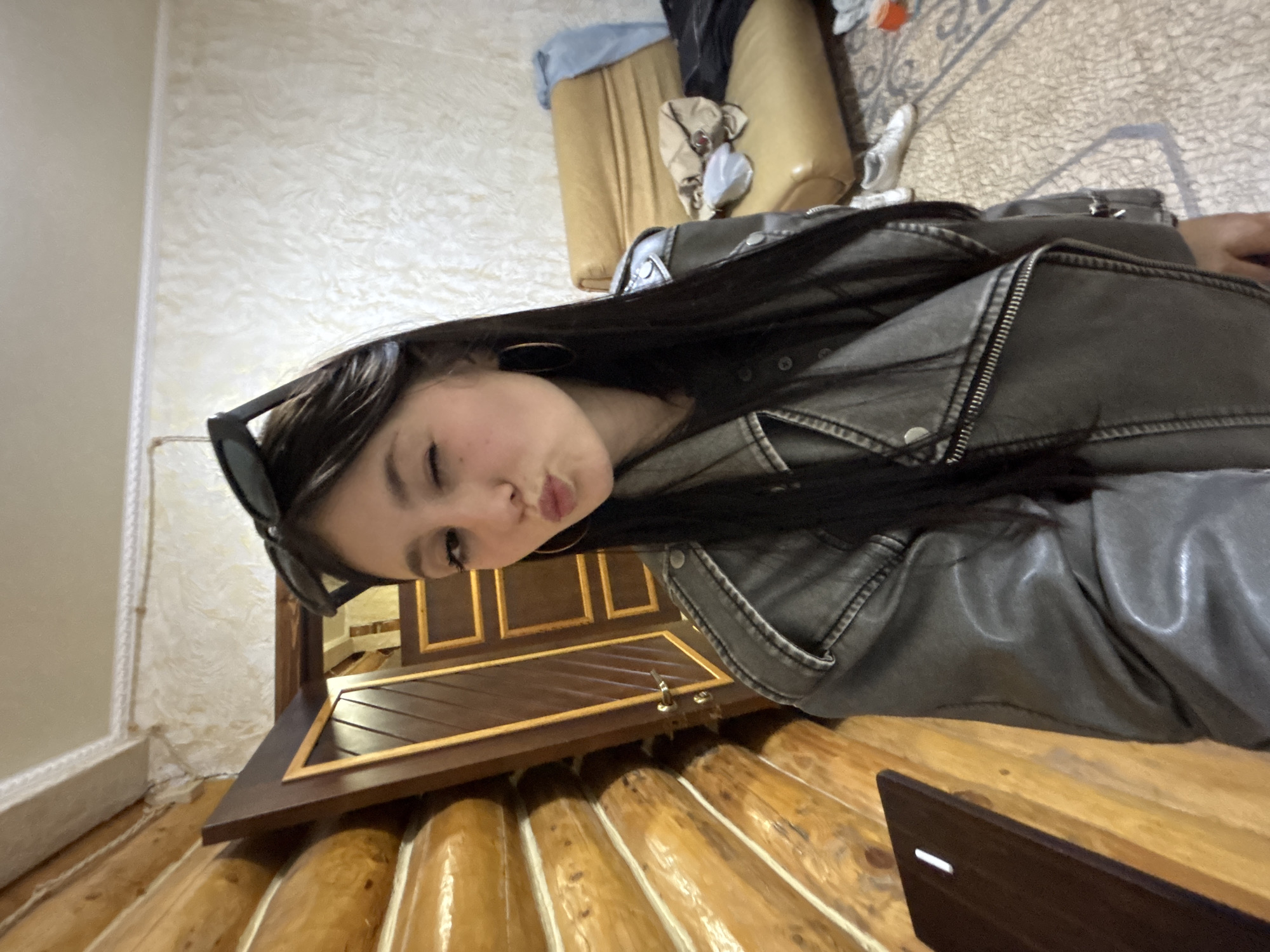A woman with long, dark hair is posing for a selfie indoors. She is wearing a gray leather jacket, hoop earrings, and sunglasses are perched atop her head. She is pouting her lips towards the camera. The background includes a light-colored, textured wall, a wooden dresser, and wooden logs. The precise location in Chaikovsky, Russia cannot be determined based on this image.