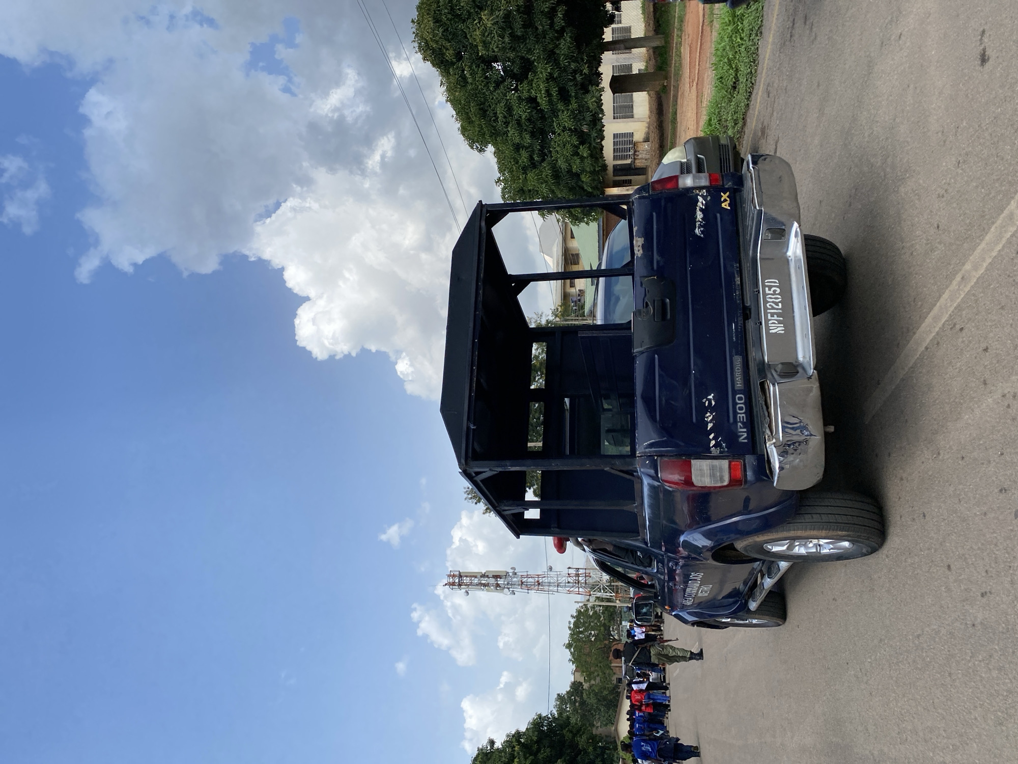 A dark blue police utility truck with a reinforced cage in the rear is parked on an asphalt road. The truck has a chrome bumper and the license plate reads "NPF1285D". Text on the side of the truck is partially visible, with "NP300 HARDBODY" and "AREA COMMANDERS" discernible. A person in military uniform stands near the truck. In the background, a telecommunications tower is visible against a bright blue sky with scattered white clouds. There are trees and buildings in the distance. A group of people, some wearing blue shirts and others red, are lined up on the side of the road, facing the truck. The scene appears to be taking place during the daytime, under fair weather conditions. The context suggests an official gathering or event possibly related to law enforcement or security operations in Jos, Nigeria.