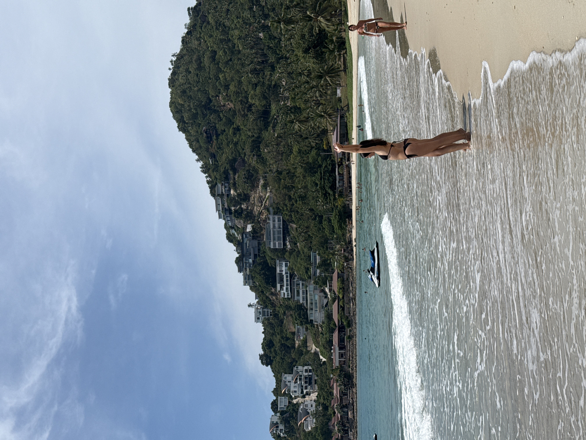 An exterior, medium shot shows a tropical beach scene with a steep, tree-covered hillside lined with white villas in the background. The ocean, with gentle waves and white foam, fills the foreground and mid-ground. A woman in a black bikini stands in the shallow water, facing away from the camera with her arms raised. Further down the beach, another woman in a black and white bikini walks along the wet sand. A blue and white jet ski is partially visible in the water near the shore. The sky is bright blue with scattered white clouds. The location is identified as a beach in the Ko Samui District of Thailand.