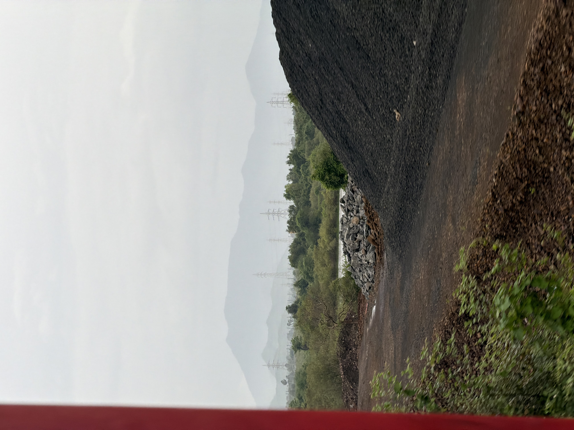 The image depicts an outdoor industrial scene in Saphale, India, with a focus on a large pile of black, granular material, likely coal or ore, dominating the right foreground. A dirt path or road runs through the middle ground, separating the large pile from a smaller pile of grey rocks and debris to its left. Further into the background, there is a body of water with green foliage and trees lining its banks. Beyond the vegetation, a hazy landscape of rolling hills or mountains is visible, punctuated by several tall electricity pylons with visible transmission lines. The sky is overcast and appears to be a pale, muted grey. A vertical red pole is partially visible on the extreme left edge of the frame, suggesting the perspective might be from a moving vehicle or a structure. The overall impression is one of an industrial area situated in a natural, somewhat undeveloped environment.