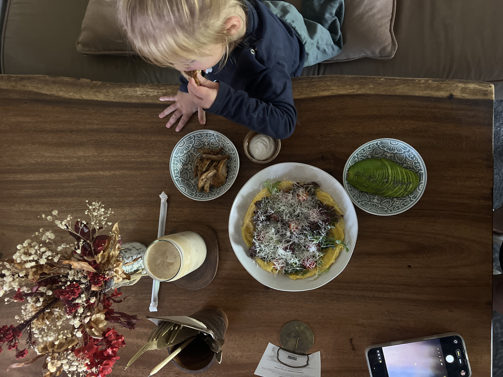 An overhead shot shows a child with blonde hair eating from a small bowl on a wooden table in North Kuta, Indonesia. The child is wearing a dark blue long-sleeved shirt and a green garment is visible on their lap. The table also holds a plate of food with shredded cheese and greens, a bowl of sliced avocado, a glass of a light-colored beverage, a small bowl with what appears to be chicken, and a small dish of white sauce. A vase of dried flowers and cutlery in a wooden holder are also present. A smartphone is visible on the left side of the table, displaying a camera interface. The background includes a glimpse of a sofa.