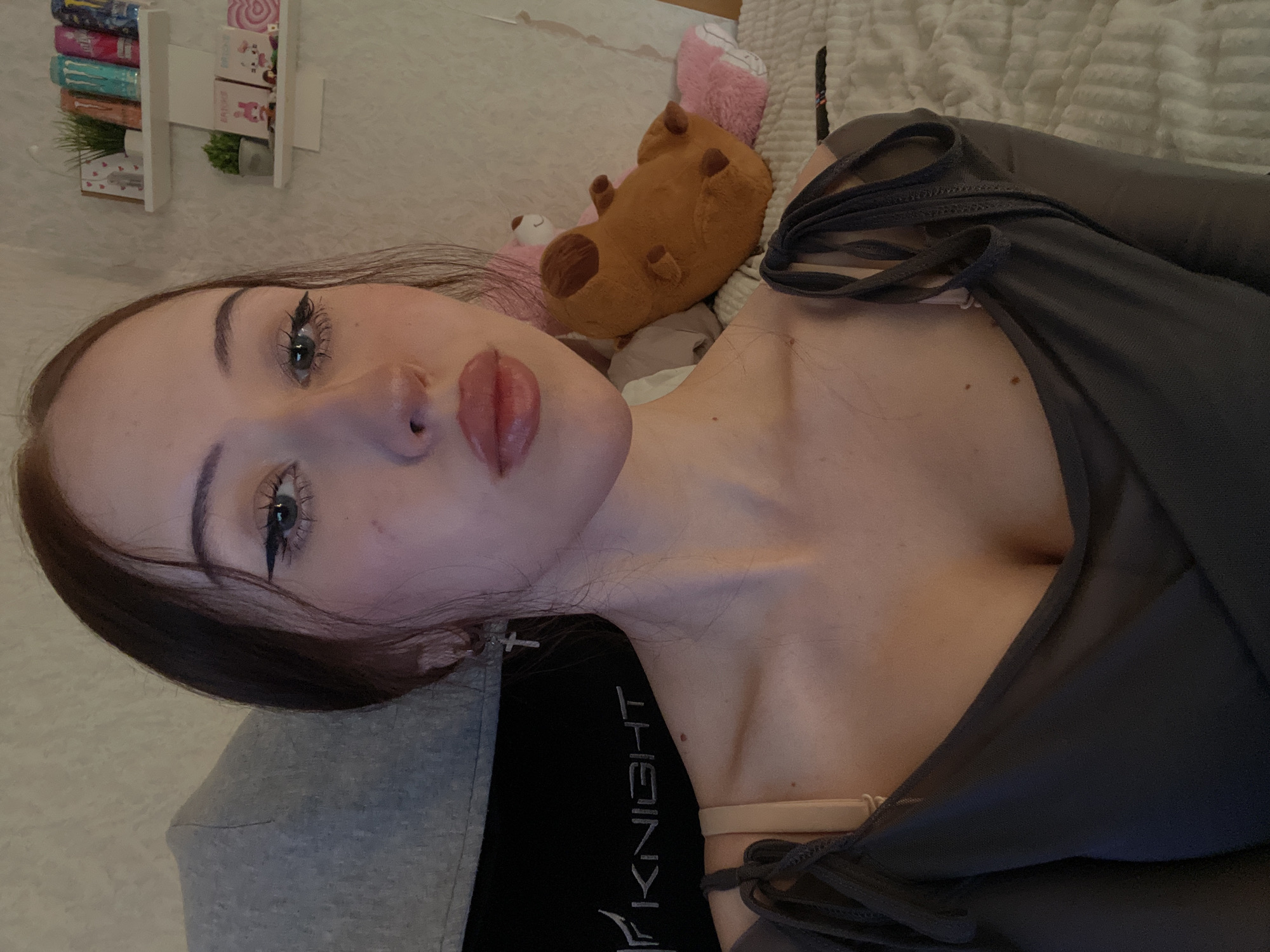 A young female, approximately early twenties, with fair skin and dark brown hair, is depicted in a medium shot looking directly at the camera. Her hair is styled to frame her face. She has prominent black winged eyeliner and mascara on her light-colored eyes. Her lips are full, glossy, and a pinkish-nude color, slightly parted. A silver-colored cross earring is visible in her left ear. She wears a dark gray, long-sleeved, v-neck top with tie-detail straps on the shoulders. A light beige bra strap is visible on her left shoulder. A small mole is present on her right cheek below her eye, and a few more are on her left collarbone area.
The setting is an indoor room, likely a bedroom. To the subject's left, a portion of a gray, high-backed chair, possibly a gaming chair, is visible. The word "KNIGHT" is printed in white lettering on the chair's black fabric. To the subject's right, a light-colored wall features two white floating shelves. The top shelf holds a small green artificial plant, two small framed pictures (one with red hearts), and a row of five colorful beverage cans, resembling energy drinks. The bottom shelf contains another small green artificial plant and four small rectangular items, appearing to be cards or small books, some with cartoon illustrations (e.g., a pink bunny, a white cat with glasses, a pink heart).
Behind the subject and to her right, a bed is visible with a light-colored, textured duvet or blanket. Several plush toys rest on the bed, including a brown, lumpy, dog-like or bear-like figure with darker patches, and a pink plush toy partially obscured behind it. The lighting is warm and ambient. There is no visual information within the image to confirm or deny its location in Votkinsk, Russia.
