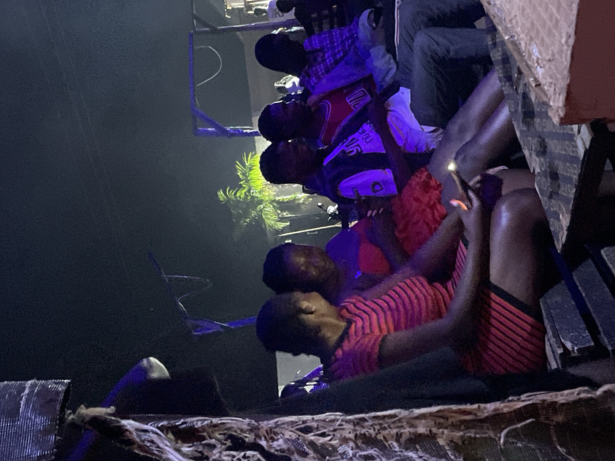 In Durumi, Nigeria, a group of people are seated outdoors at night, illuminated by ambient light. On the left, a woman wears a red and black striped dress. Next to her, another woman in a red top is visible. To their right, three men are seated, one in a white t-shirt and black vest, another in a red basketball-style jersey, and the third in a plaid shirt. The individuals appear to be engaged in an activity or watching something. The background shows a dimly lit environment with some foliage and structures suggesting an outdoor gathering or event. The overall scene is dark, with artificial lighting casting shadows and highlighting some figures.