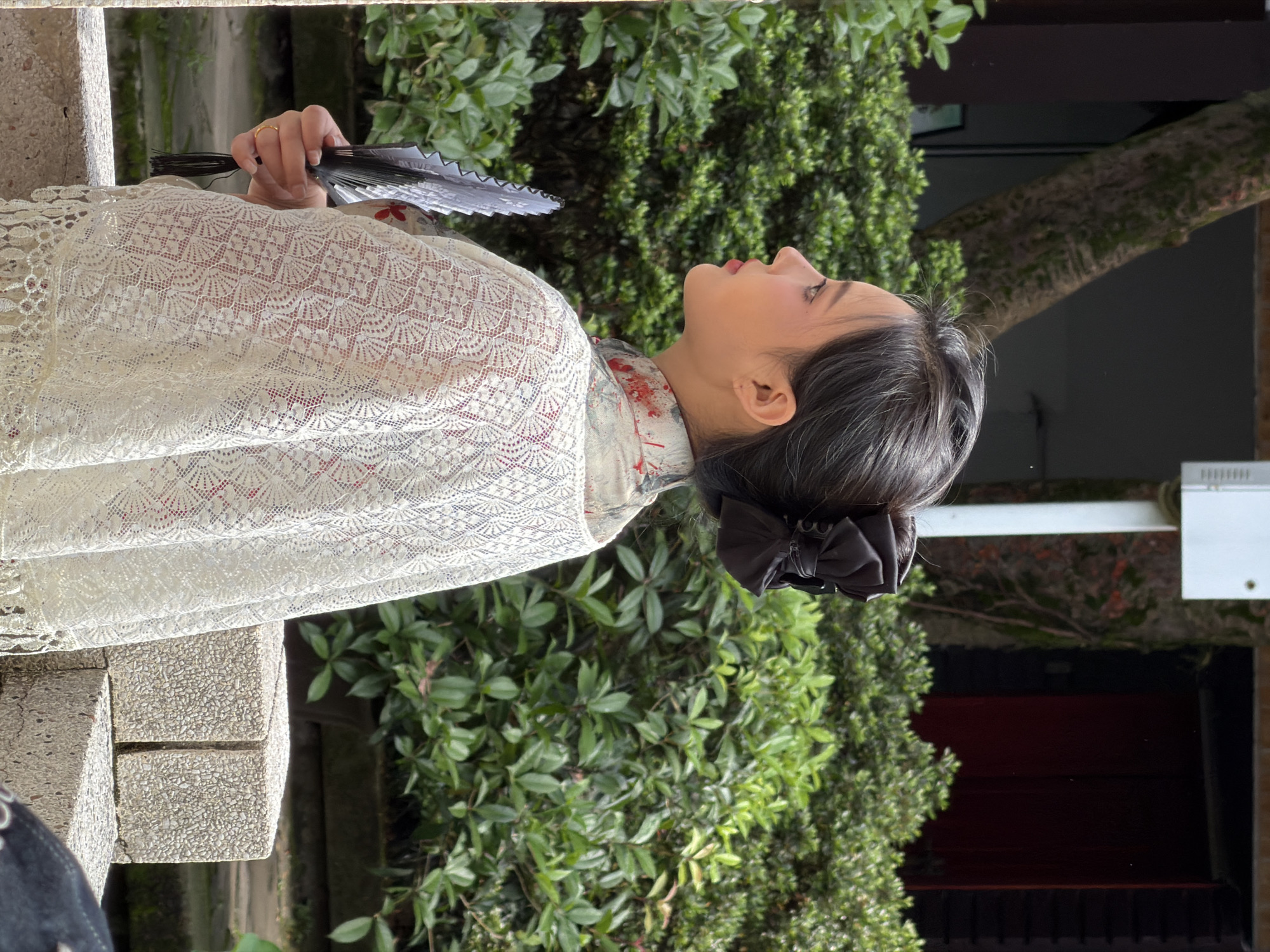 The image shows a young East Asian woman from the chest up, seen in profile facing left, standing outdoors. She has dark hair pulled back in a bun adorned with a large, dark bow. She is wearing a white, lace-like garment over a red undergarment with a floral pattern visible at the collar. In her right hand, she holds a black folding fan. The background consists of lush green foliage and a blurred building structure. The scene appears to be a garden or courtyard, possibly in a park or a residential area within Yuzhong District, China. The lighting suggests it is daytime, with soft shadows indicating overcast conditions or diffused sunlight.