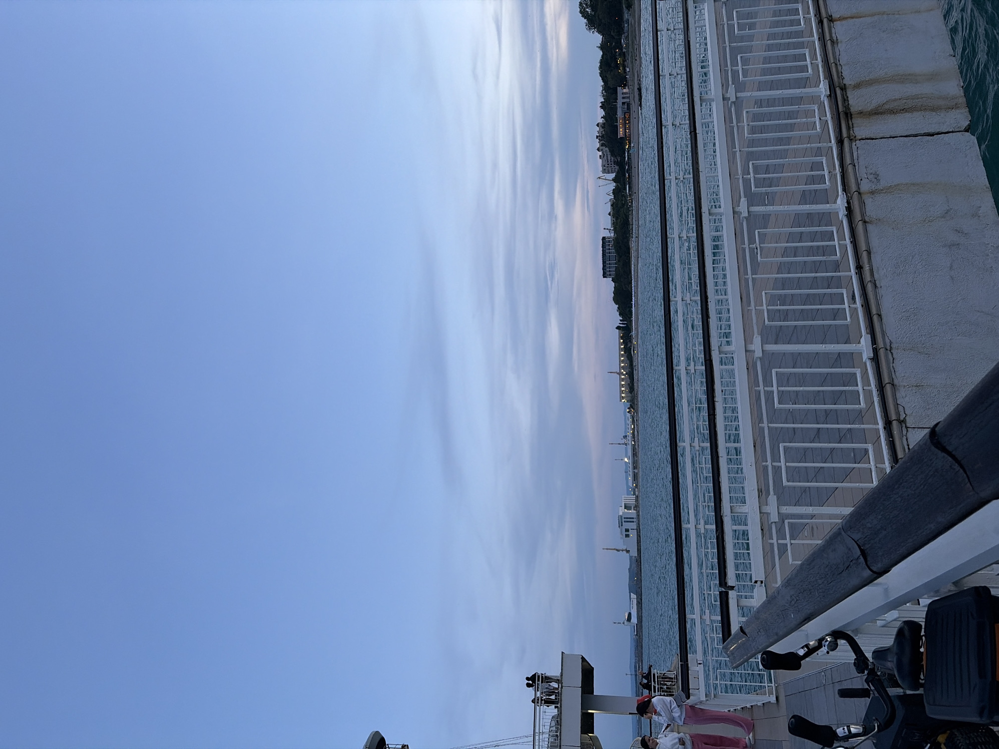The image captures a scene at the Burgas Sea Station in Burgas, Bulgaria. In the foreground, a bicycle is parked, with its handlebars and part of its frame visible. A white railing with a dark wooden handrail runs along the edge of the walkway. A portion of the concrete pier structure is also visible, showing some staining.
In the mid-ground, a white railing lines a pedestrian walkway paved with grey rectangular tiles. Beyond the walkway, the sea is visible, with gentle waves. Several structures are located on the opposite shore, including what appears to be a ferry or ship, white buildings, and some cranes. The sky above is a soft blue with wispy clouds, suggesting it is late afternoon or early evening.
In the left mid-ground, three people are standing on an elevated platform, silhouetted against the sky. Further to the left, two young women wearing light-colored hoodies and pink pants are standing on the walkway, with one looking towards the sea and the other looking down.