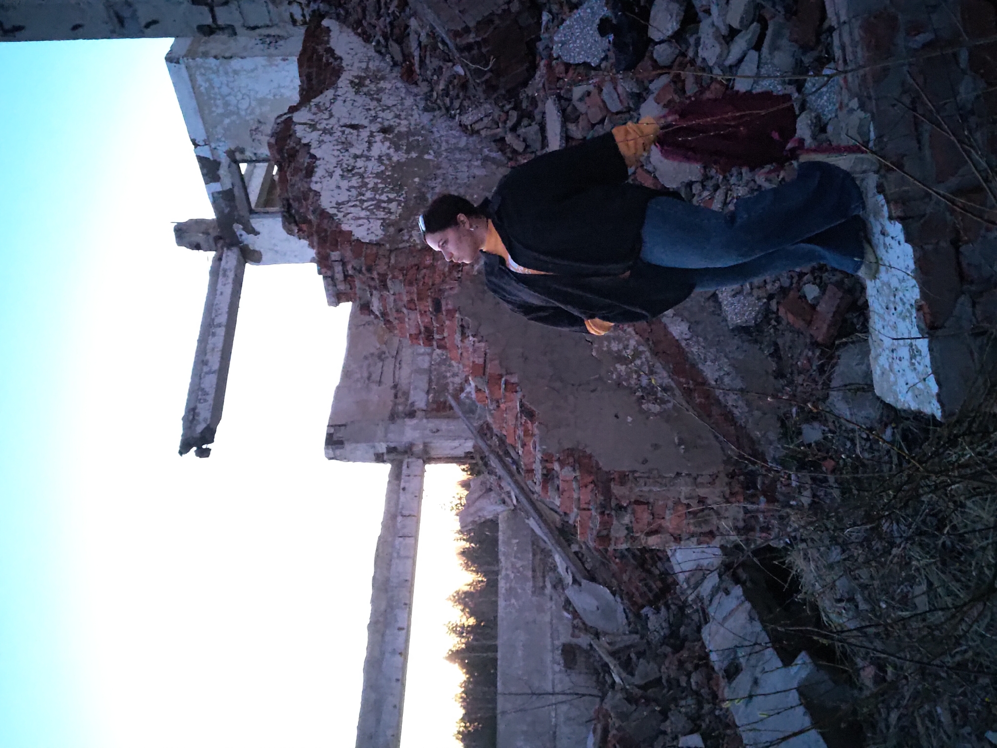 The image depicts a scene in what appears to be the ruins of a building in Марковский, Russia. The structure is heavily damaged, with exposed brick and concrete. A person is standing amidst the rubble, wearing a dark jacket and jeans. The sky is visible through the skeletal remains of the building, suggesting an outdoor location. Debris and vegetation are scattered throughout the scene.