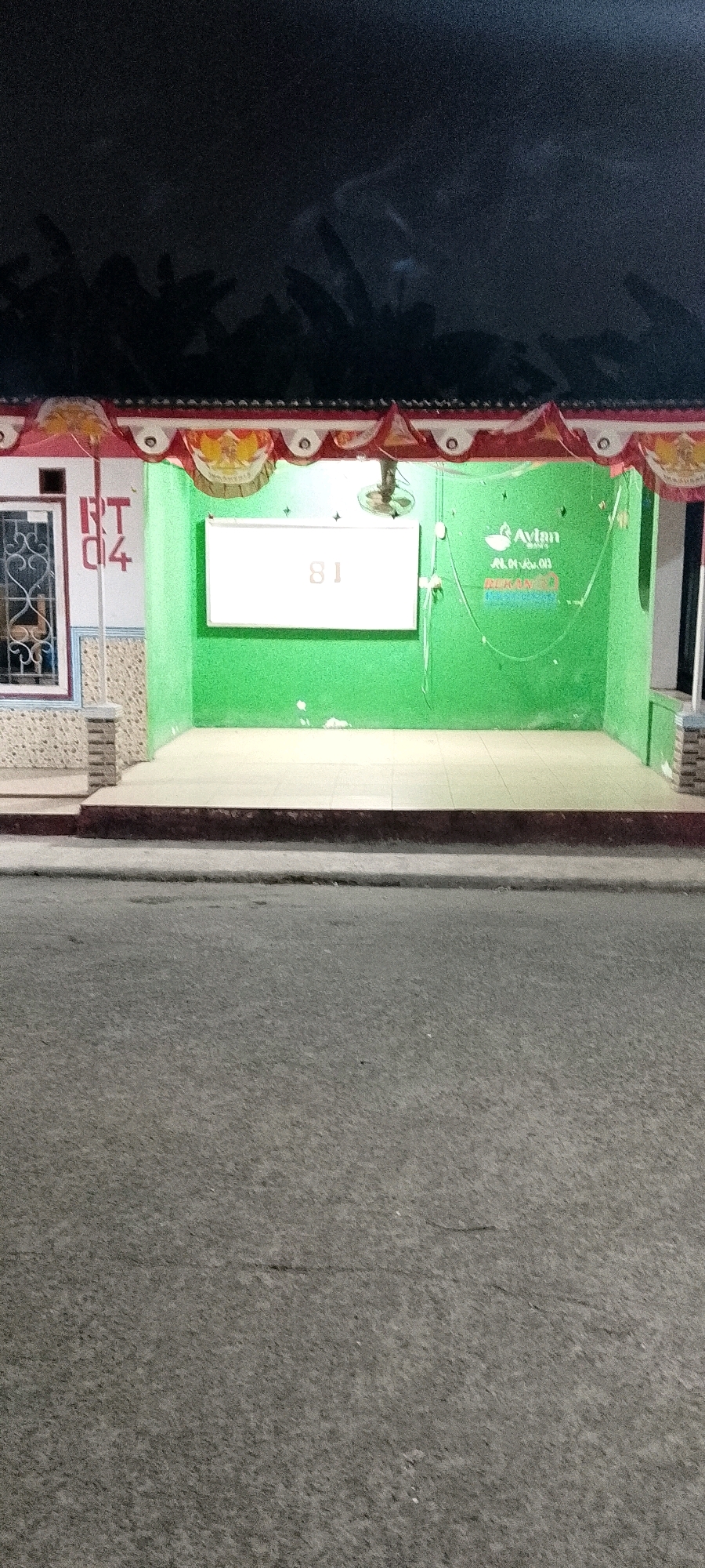 The image shows a night view of a green bus stop in Satriamekar, Indonesia. The bus stop has a white board with the number "81" on it, and the brand logo "Avlan" is visible on the green wall. The bus stop is decorated with red and white banners. The street in front of the bus stop is paved with asphalt. The sky above is dark, with some shadows of trees visible.