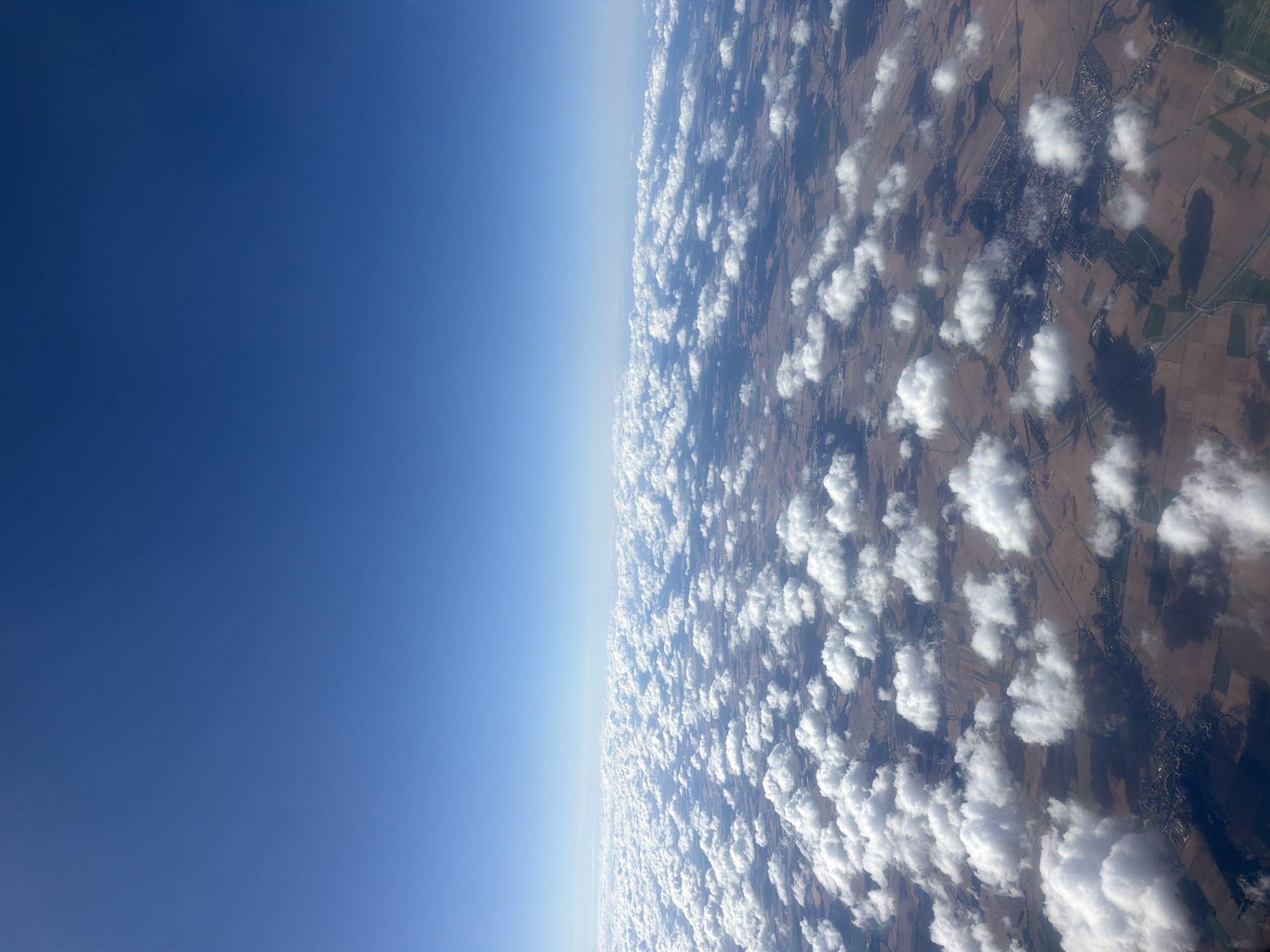 The image is an aerial view of the city of Raczkowa, Poland, as seen from an airplane. The scene is dominated by a vast expanse of white, puffy cumulus clouds scattered across a clear blue sky. Below the clouds, the landscape of Raczkowa is visible, characterized by a patchwork of brown and green fields, indicating agricultural activity. Several roads and what appear to be clusters of buildings, suggesting urban or semi-urban areas, can be discerned through the breaks in the clouds. The sun is casting shadows from the clouds onto the ground, creating a dappled light effect. No people or specific actions are discernible from this high-altitude perspective.