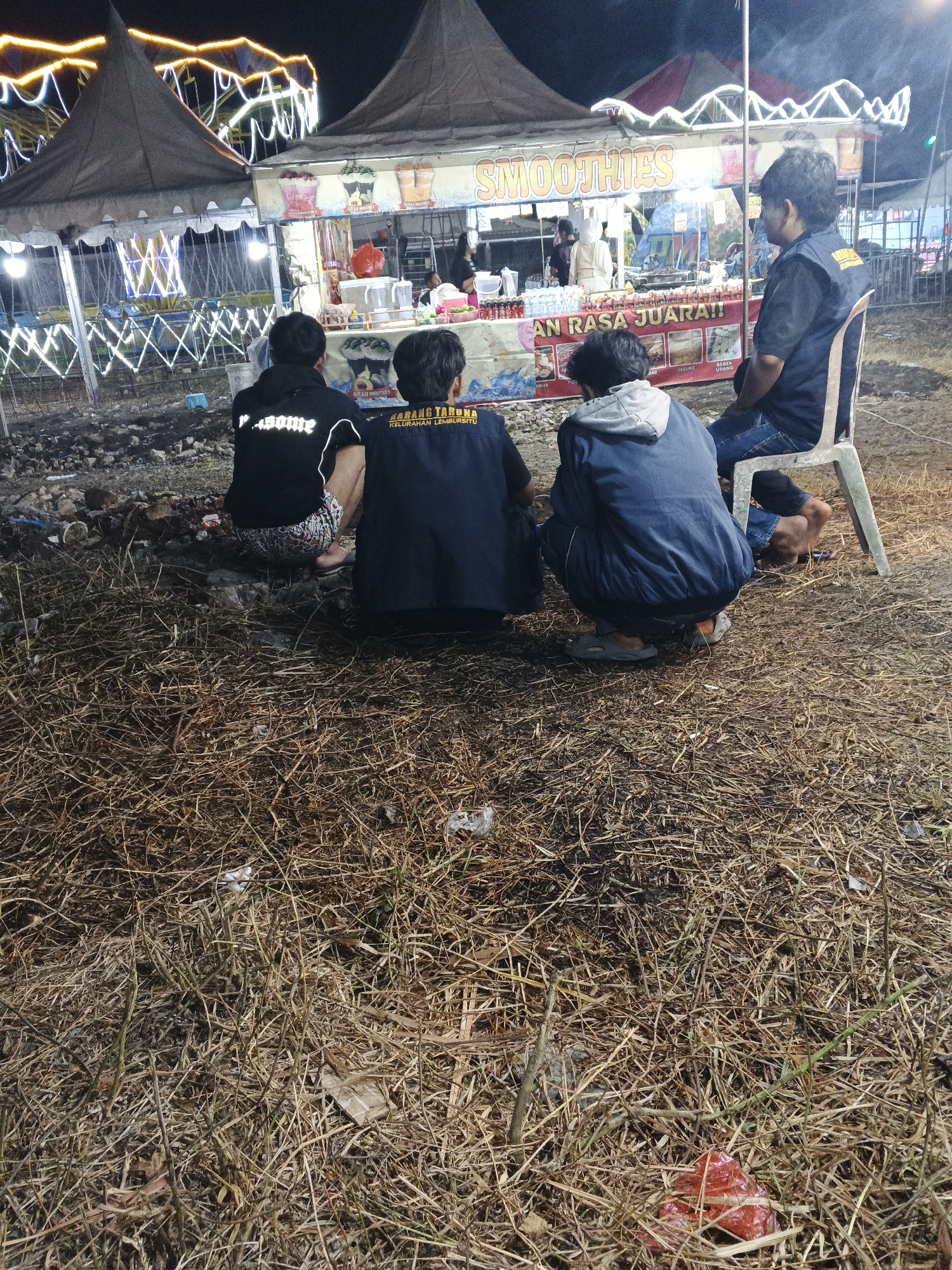 Four individuals are seen from behind, in the foreground, observing a food stall at night. The stall, identified by signage as selling "SMOOTHIES," is illuminated by strings of lights and appears to be part of an outdoor event. The ground is covered in dry grass and debris. One person is seated on a chair, while the other three are squatting or sitting on the ground. The overall scene suggests a casual gathering at a night market or fair in Sukabumi, Indonesia.